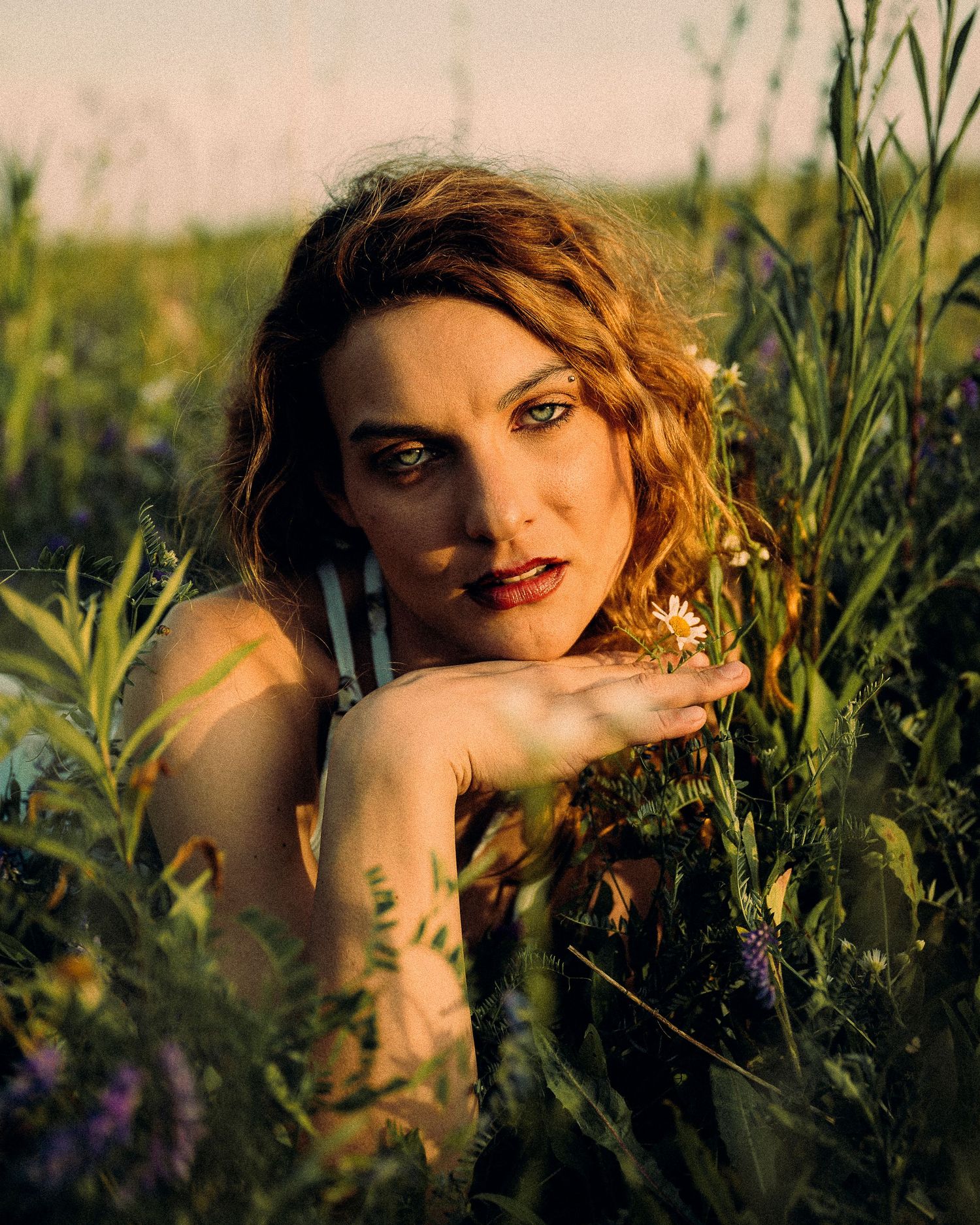 Red-haired woman with striking green eyes poses among wildflowers holding a daisy at sunset.