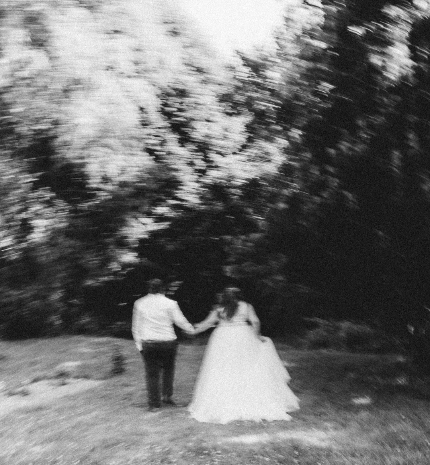Blurry black and white photo of a bride and groom holding hands while walking in nature.