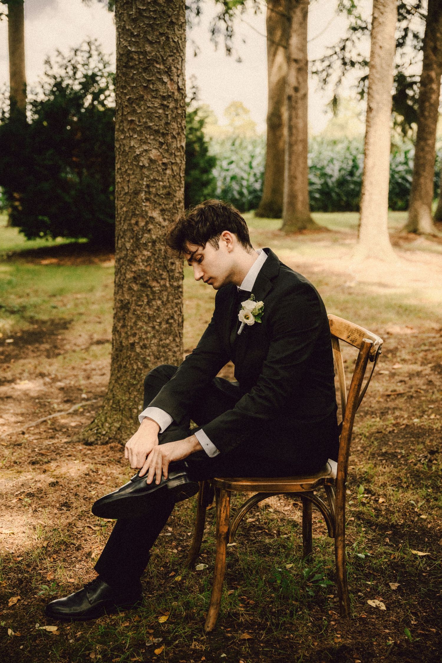 Groom in black tuxedo sits on wooden chair among trees, adjusting his shoe before the wedding ceremony.