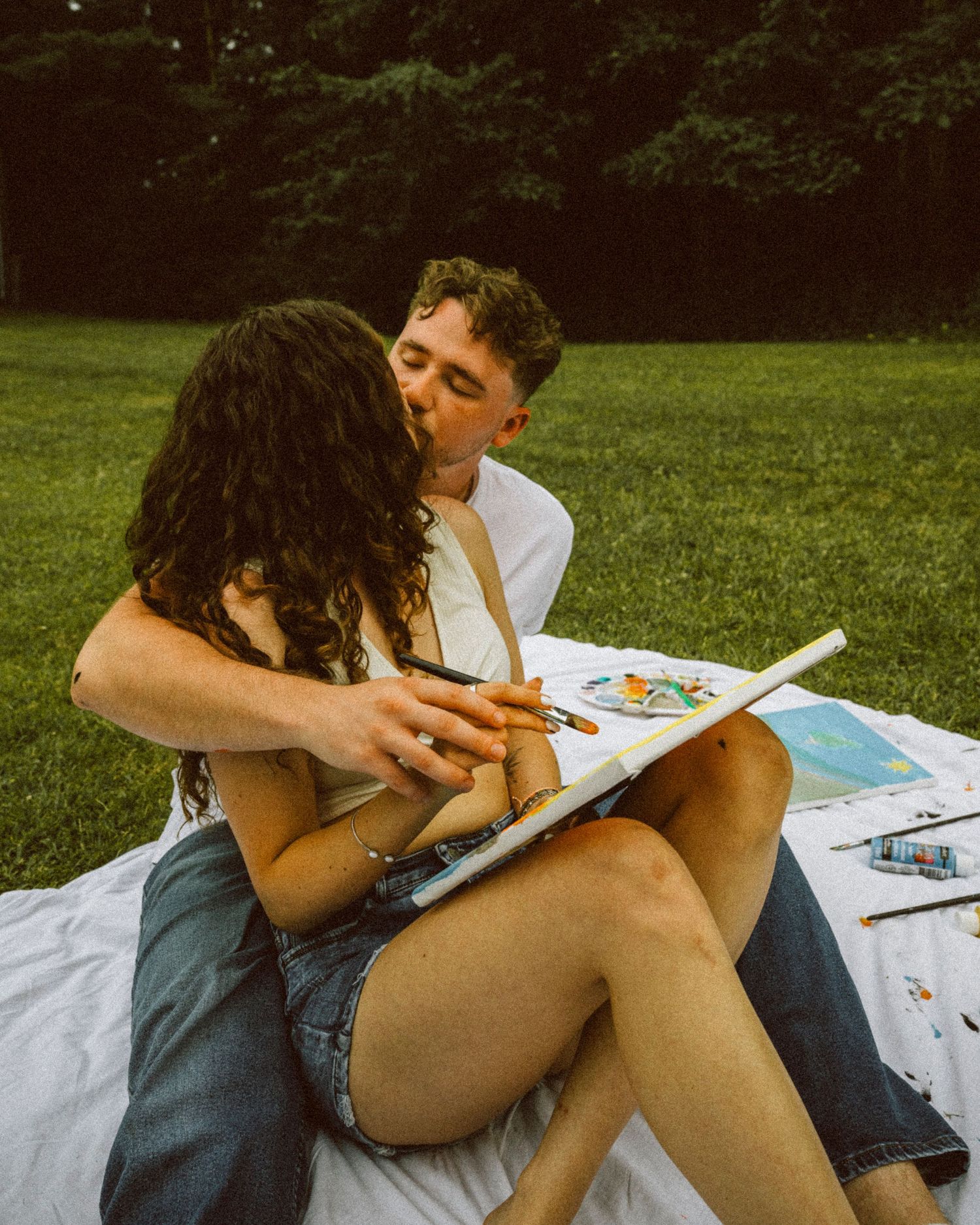 Couple sharing a kiss while sitting together on blanket holding paintbrush and canvas outdoors.