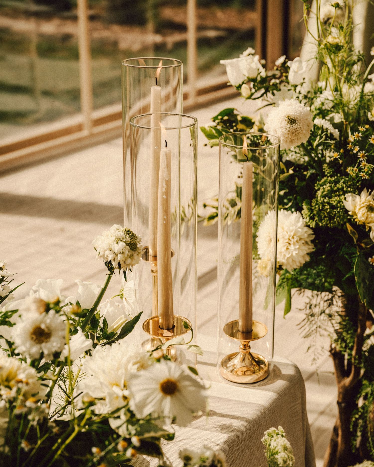 Elegant wedding decor with tall gold candlesticks in glass hurricanes surrounded by white dahlias and greenery.