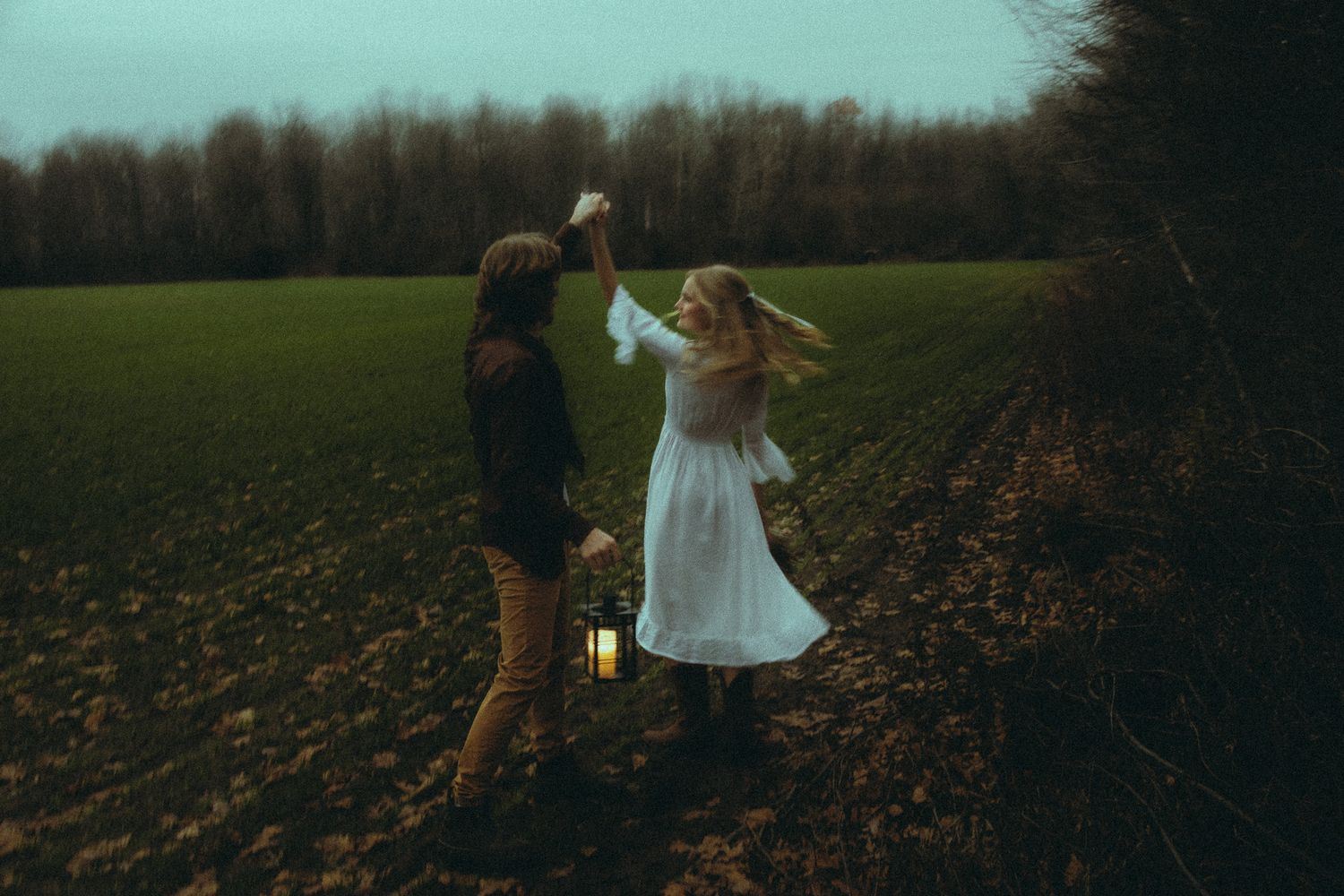 Couple dancing at dusk in a field holding a lantern, woman in white dress twirling in autumn.