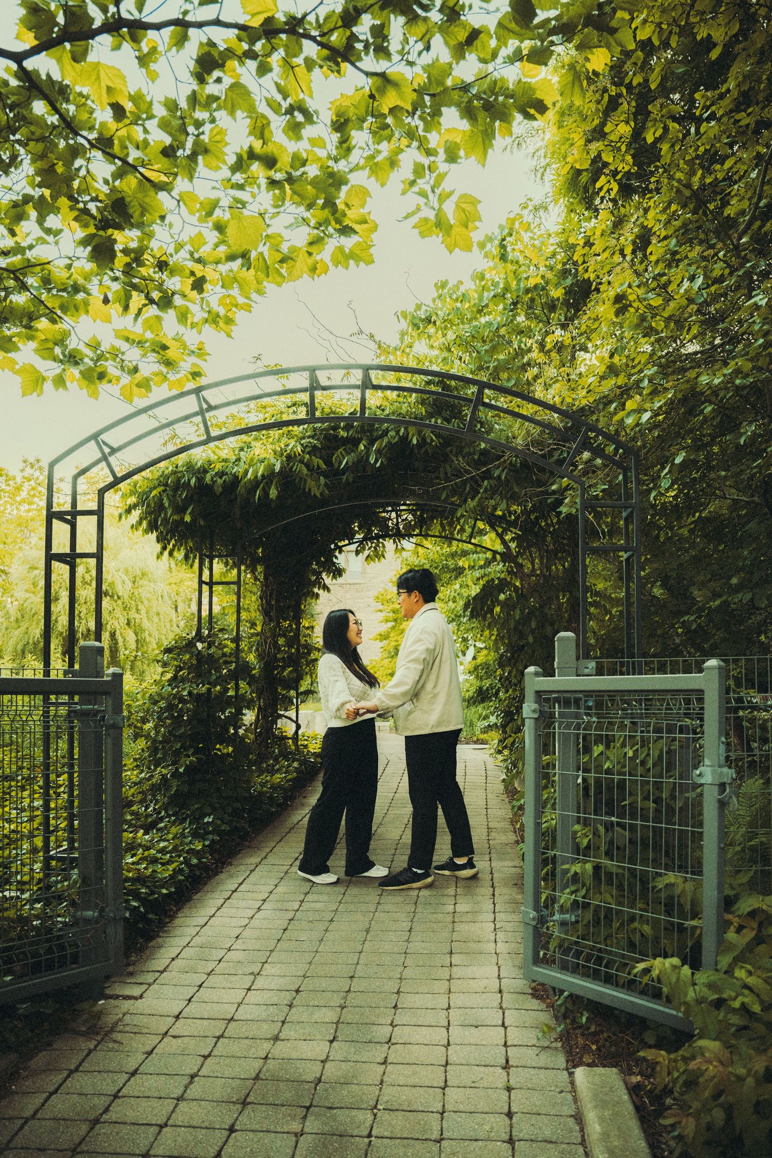 Couple stands face to face on a garden pathway beneath a lush green arched trellis.