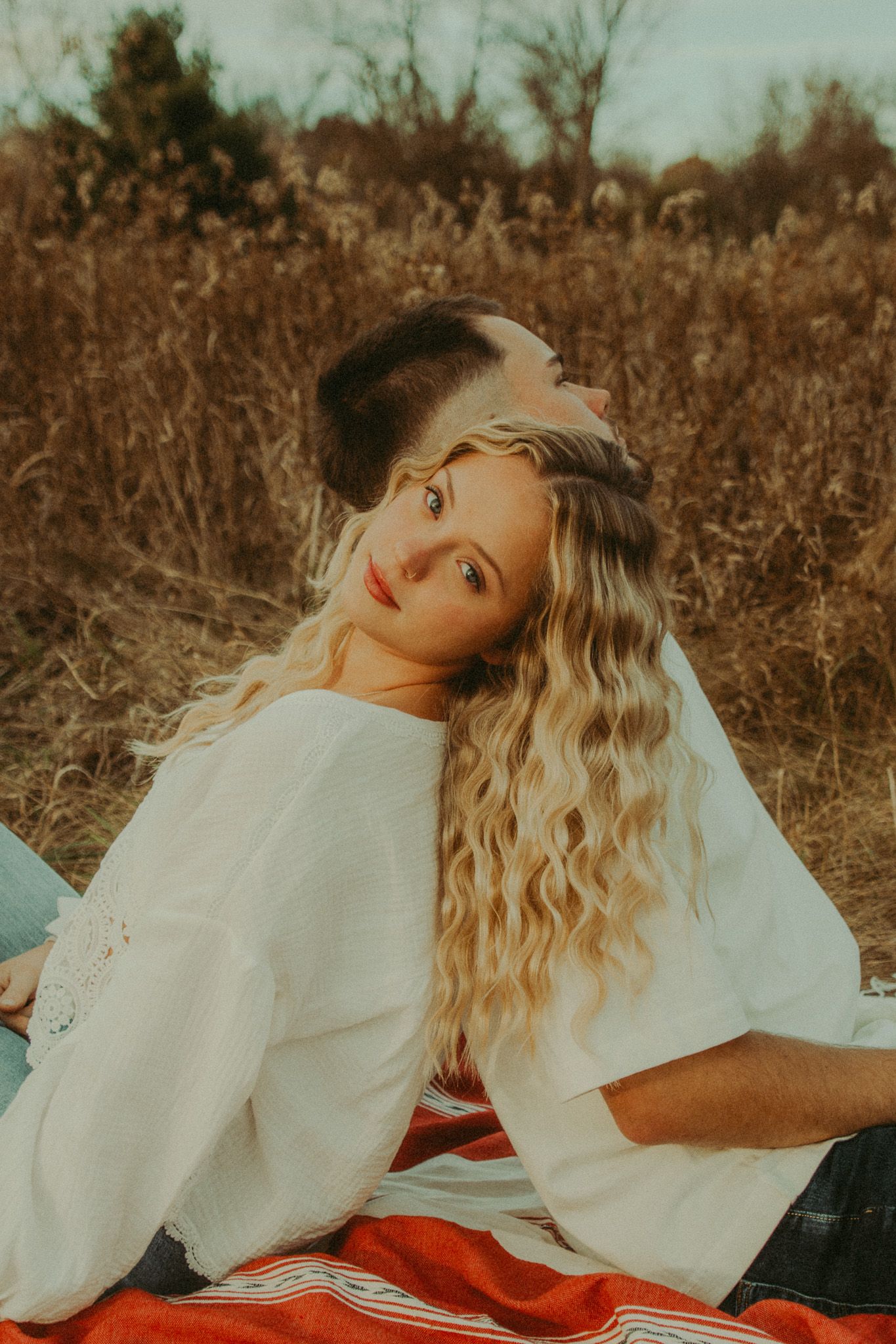 Blonde woman leaning back on man sitting together on blanket in dry autumn field at sunset.