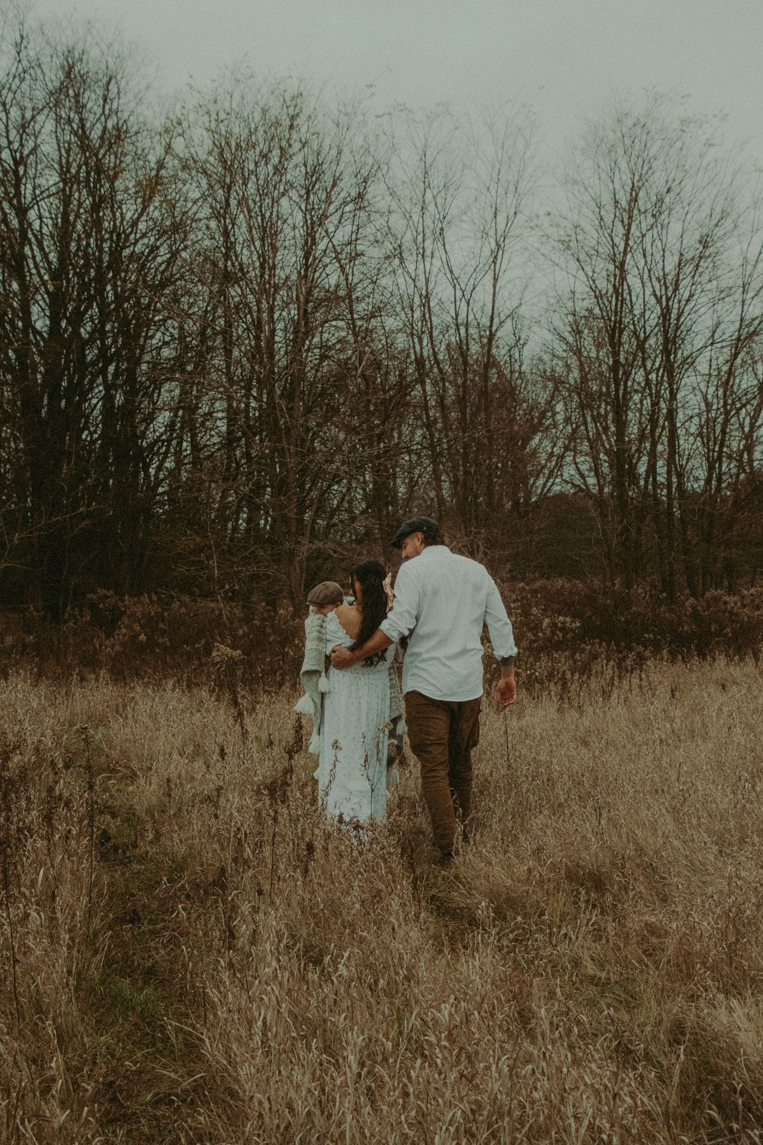 Family of three walking hand in hand through dry autumn meadow with bare trees in background.