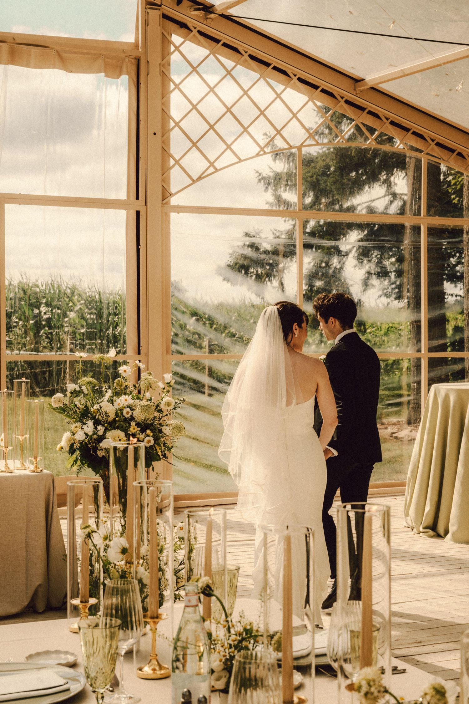 Bride and groom share intimate moment in elegant glass greenhouse wedding venue with tall candles and floral decor.