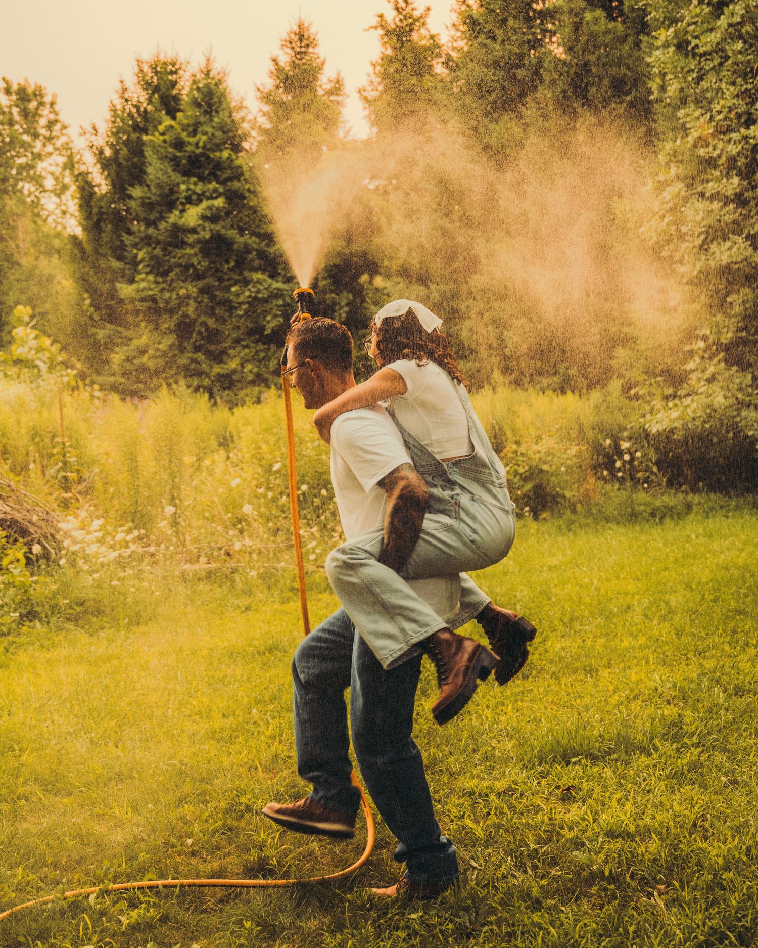 A man gives a woman a piggyback ride near a garden sprinkler in golden light.