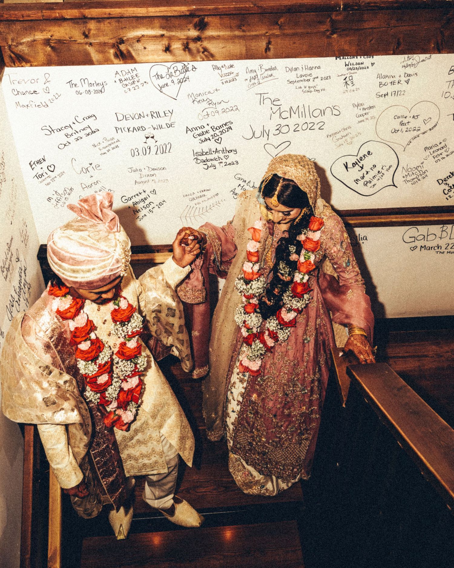 Indian bride and groom in traditional attire sign wedding guest wall on staircase.