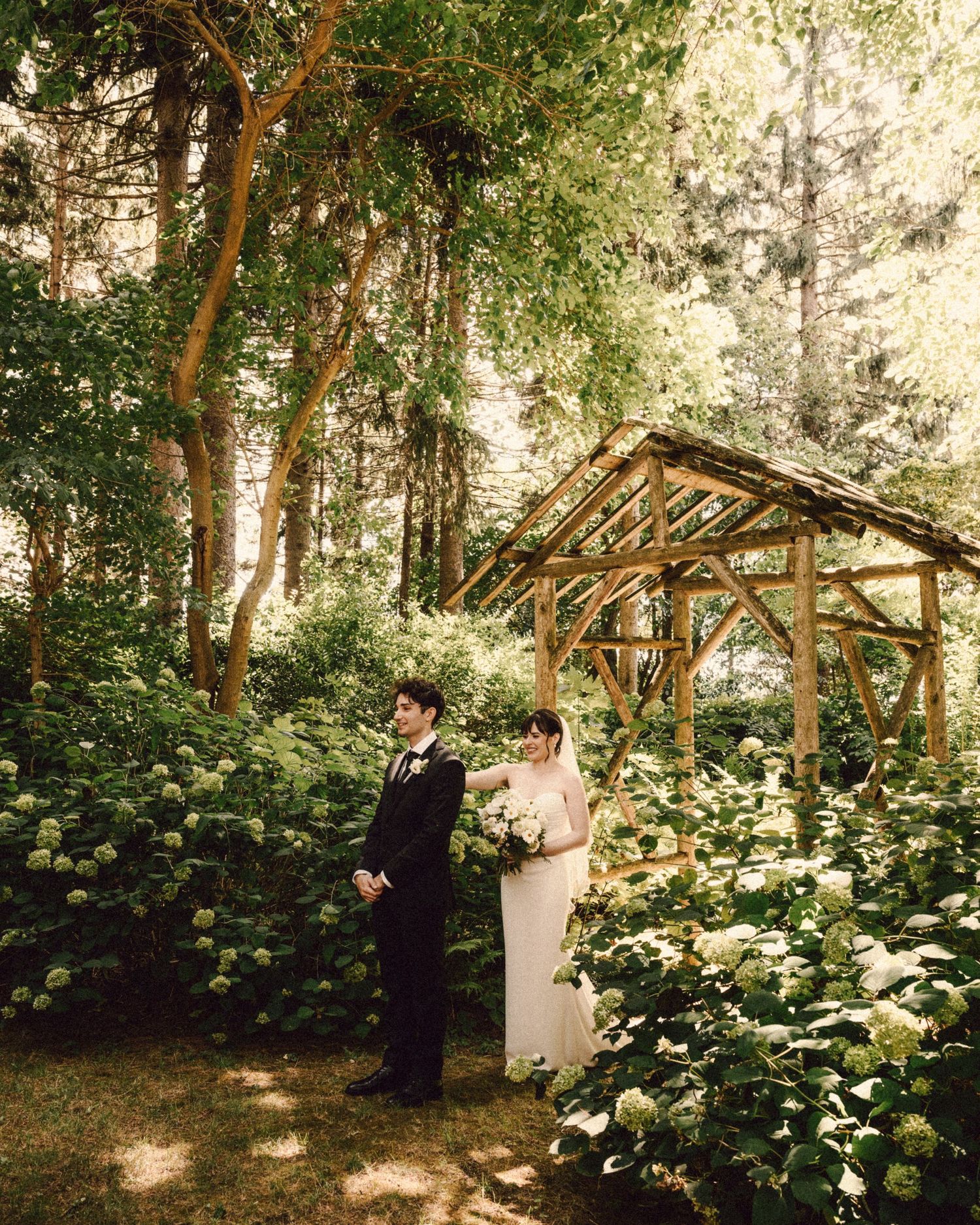 Bride and groom share a romantic first look moment beside a rustic wooden arbor surrounded by hydrangeas.