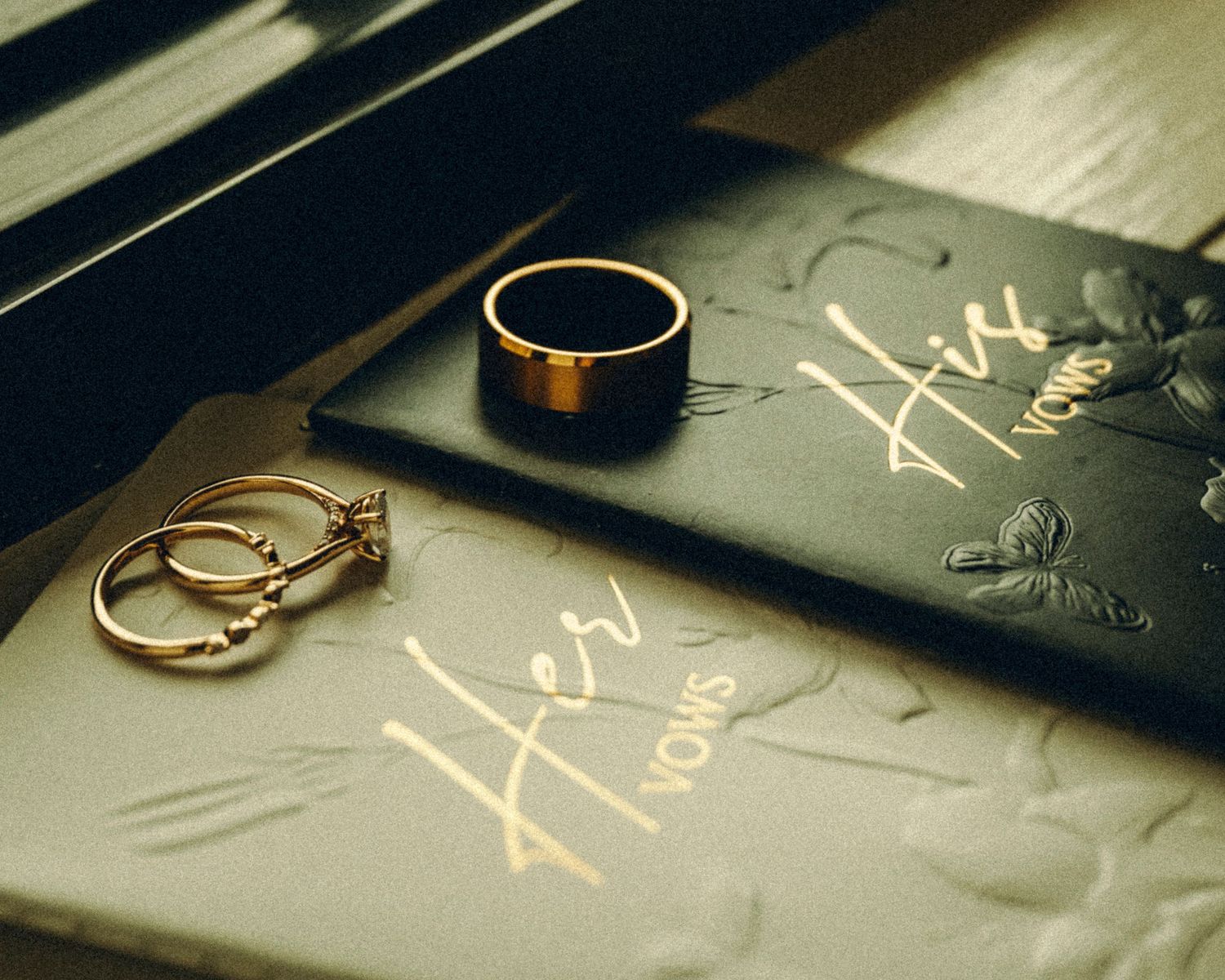 Gold wedding rings and diamond engagement ring resting on His and Her vow books in warm light.