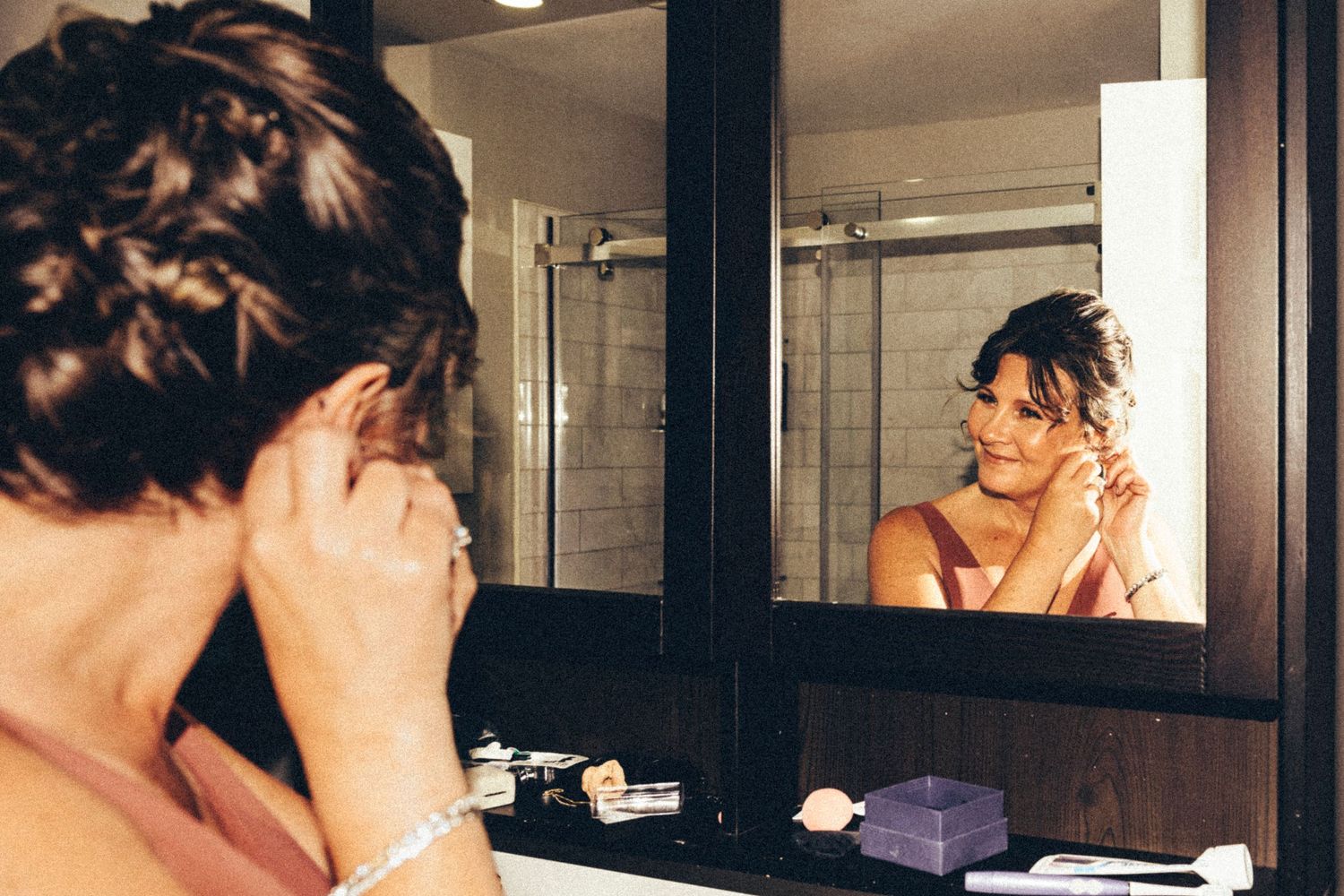 Bride smiling at reflection in bathroom mirror while putting on earrings before wedding ceremony.