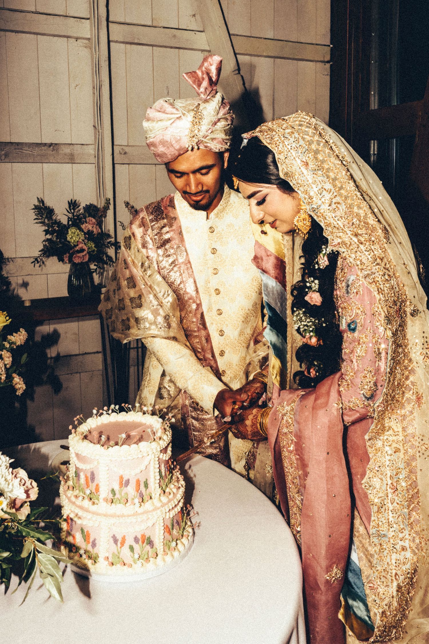 South Asian bride and groom in ornate traditional attire cut their decorated wedding cake together.