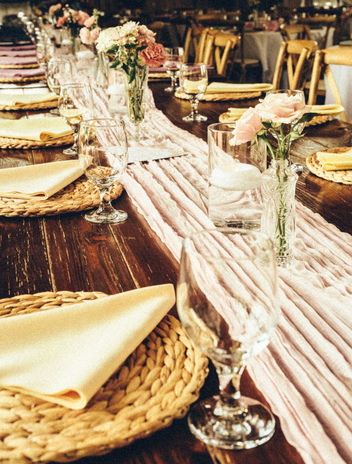Elegant wedding reception table with woven placemats, gold napkins, crystal glasses, and pink floral centerpieces on a rustic wood table.