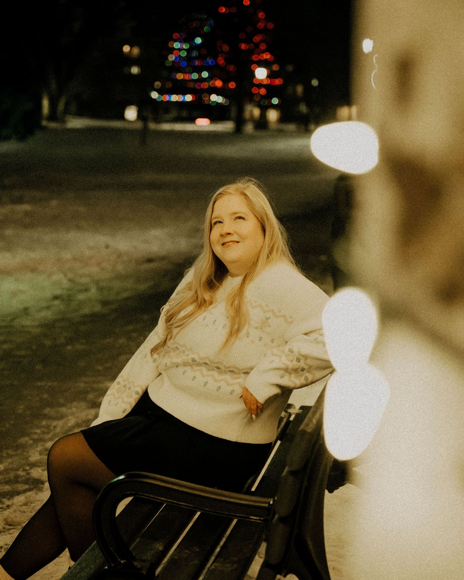 Smiling blonde woman sitting on a bench surrounded by colorful holiday lights.