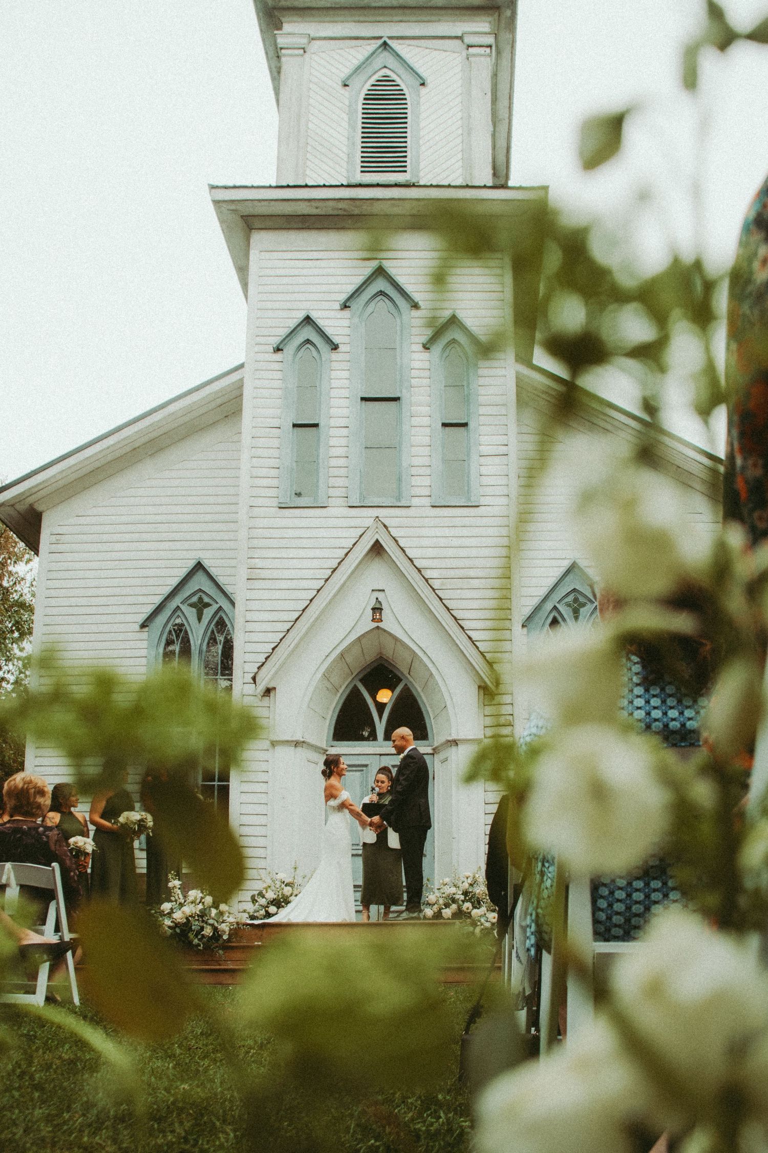 Outdoor wedding ceremony at a charming white chapel with Gothic arched windows and floral decor.