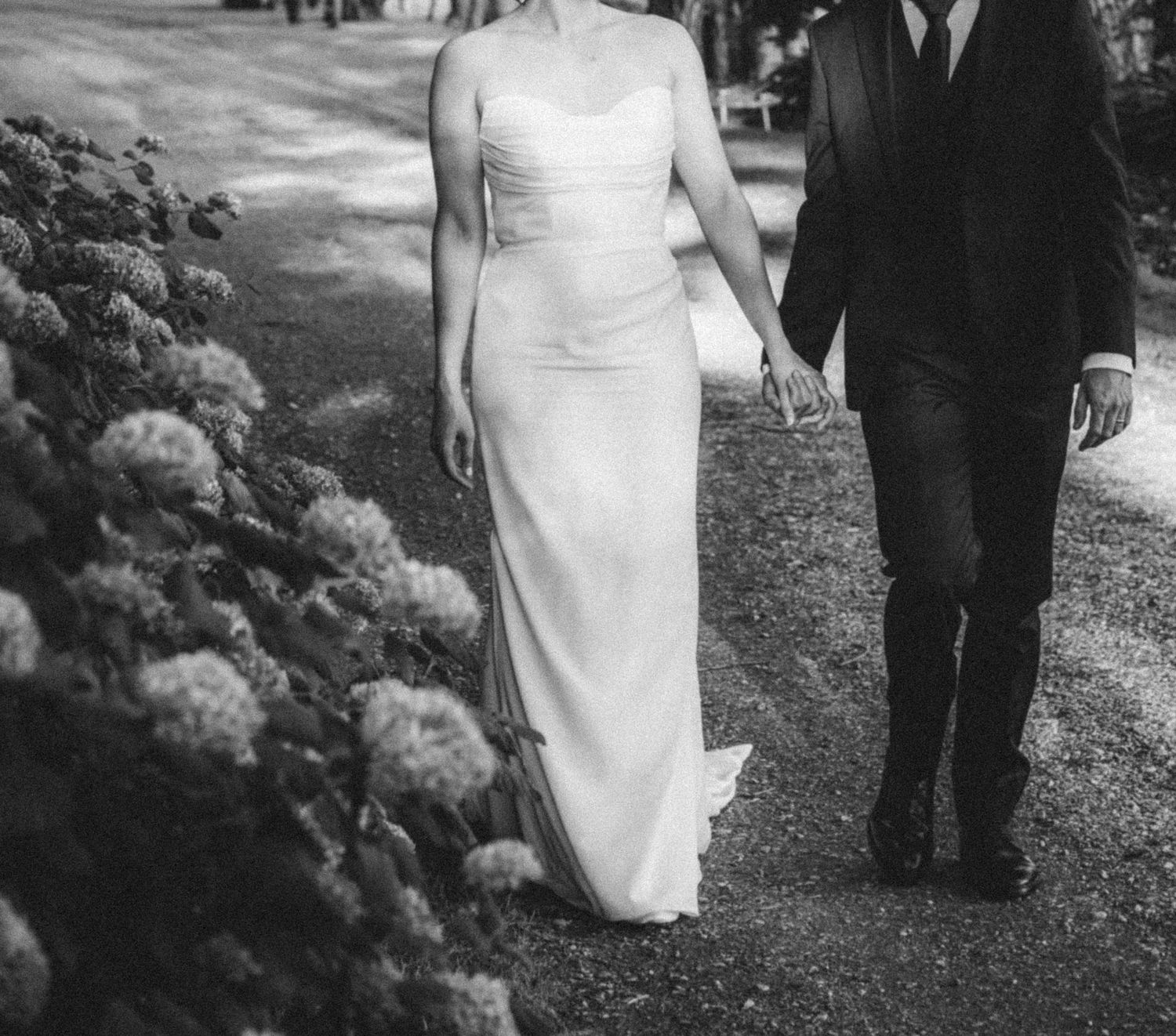 Bride in elegant white gown walking with groom past hydrangeas.