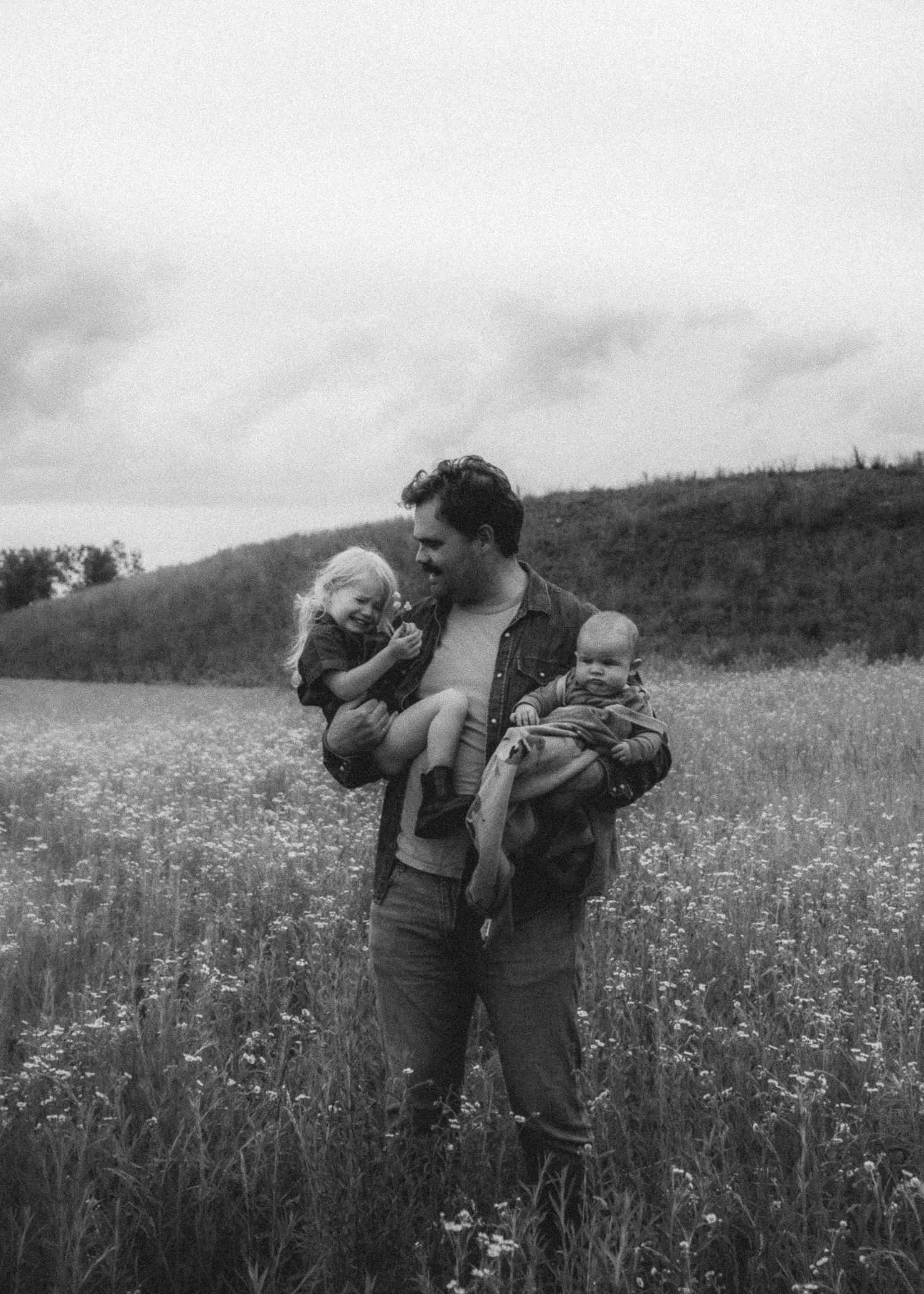 Black and white photo of father holding toddler and baby in blooming meadow with rolling hills behind.