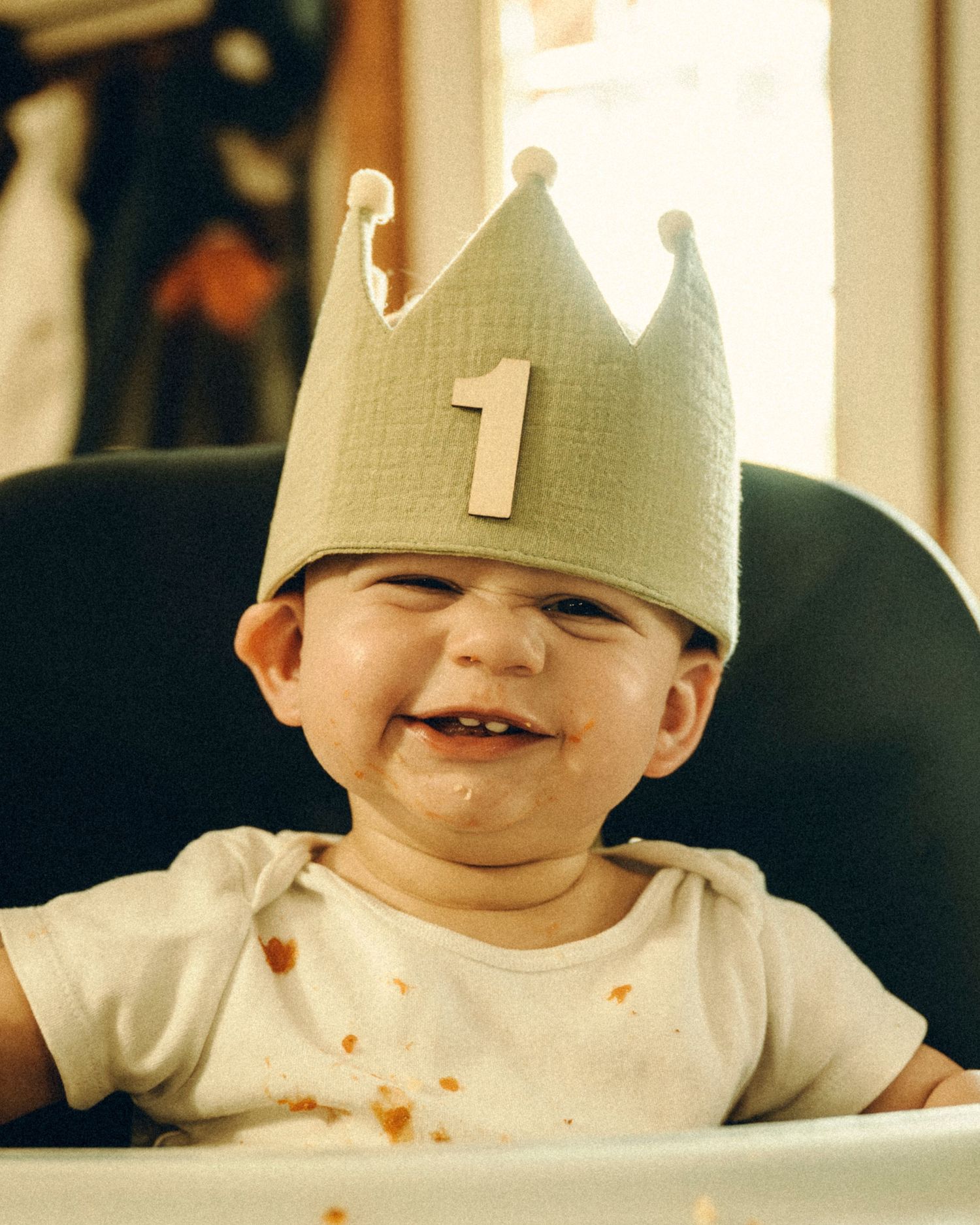 Smiling one-year-old baby wearing a crown hat with number 1 sitting in high chair.