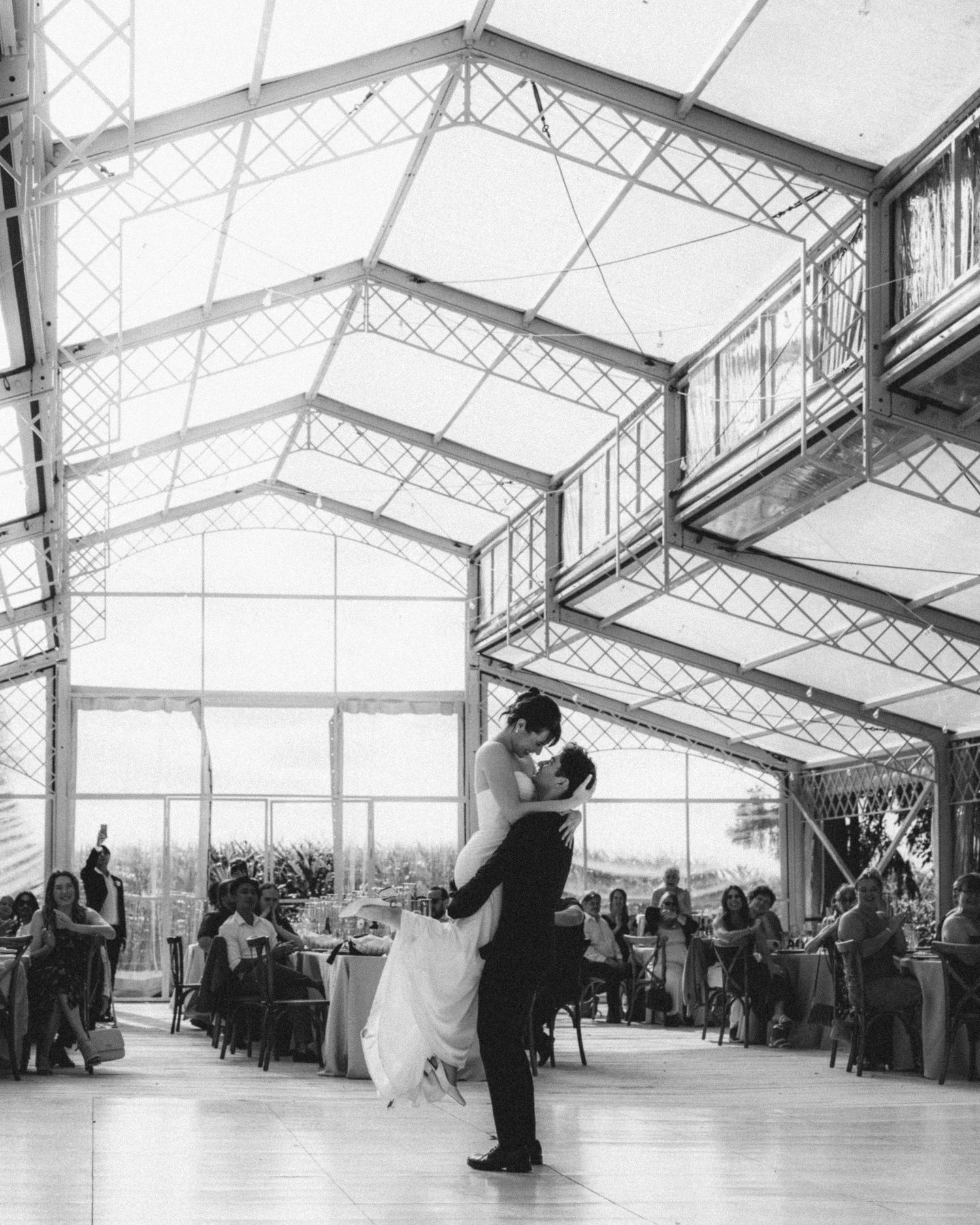 Black and white photo of groom lifting bride during first dance in stunning glass greenhouse wedding venue.