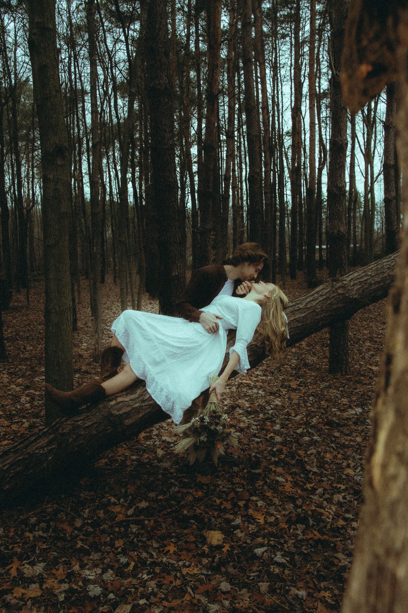 Romantic couple kissing on a fallen log in a dark autumn forest, woman in white dress.