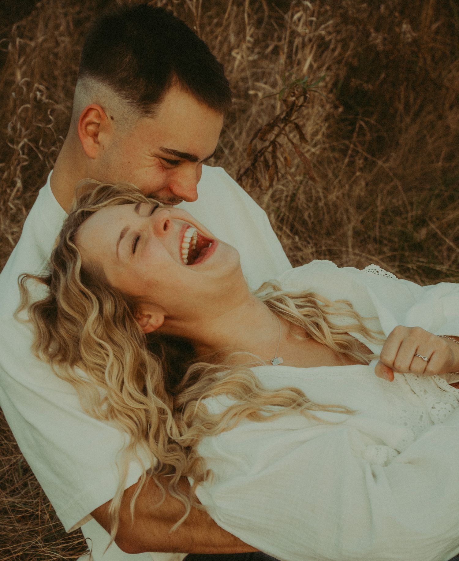 Couple laughing and playing together lying in dry grass field during golden hour.