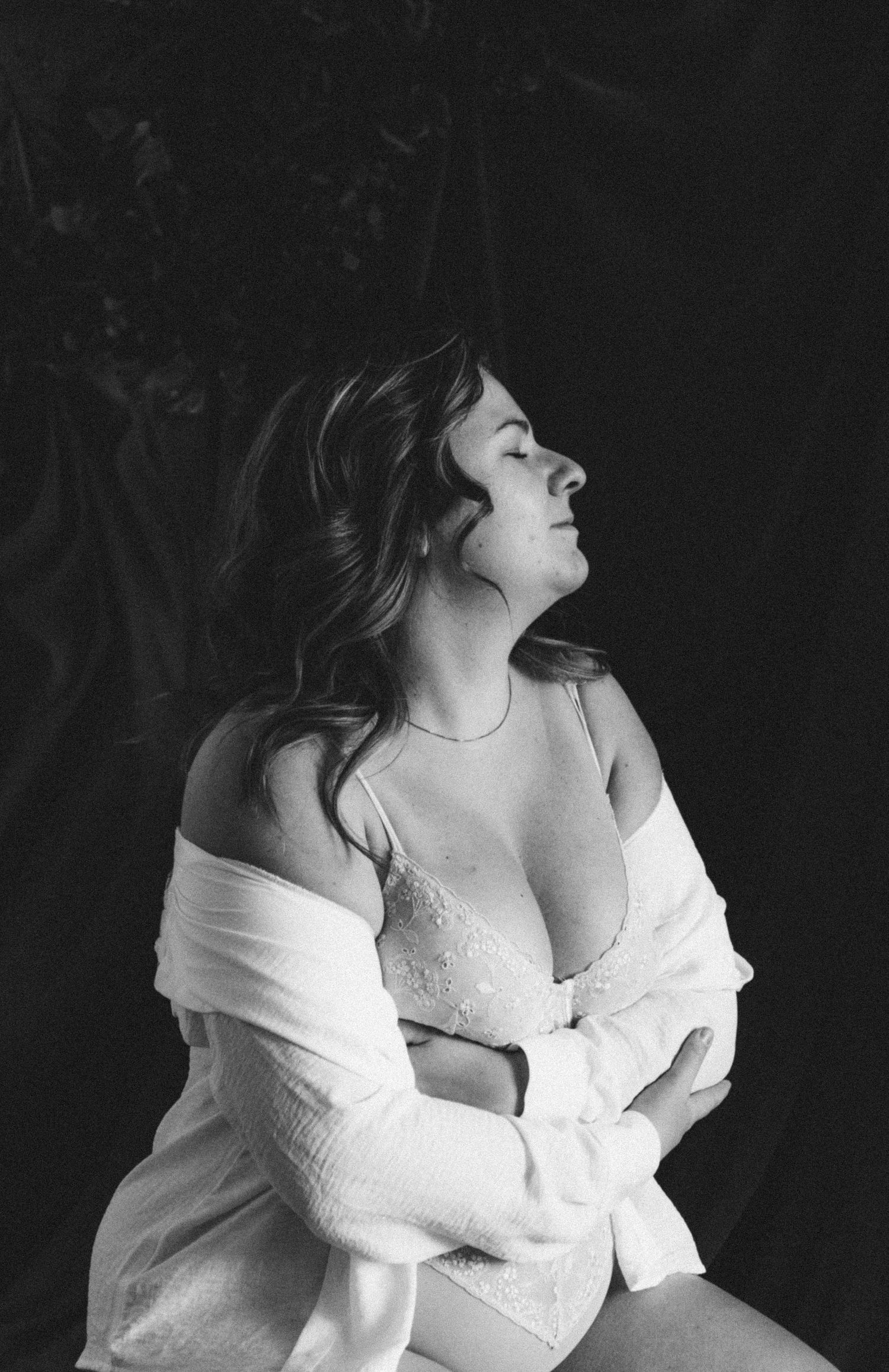 Black and white boudoir portrait of woman in lace lingerie with open white shirt, eyes closed, dark background.