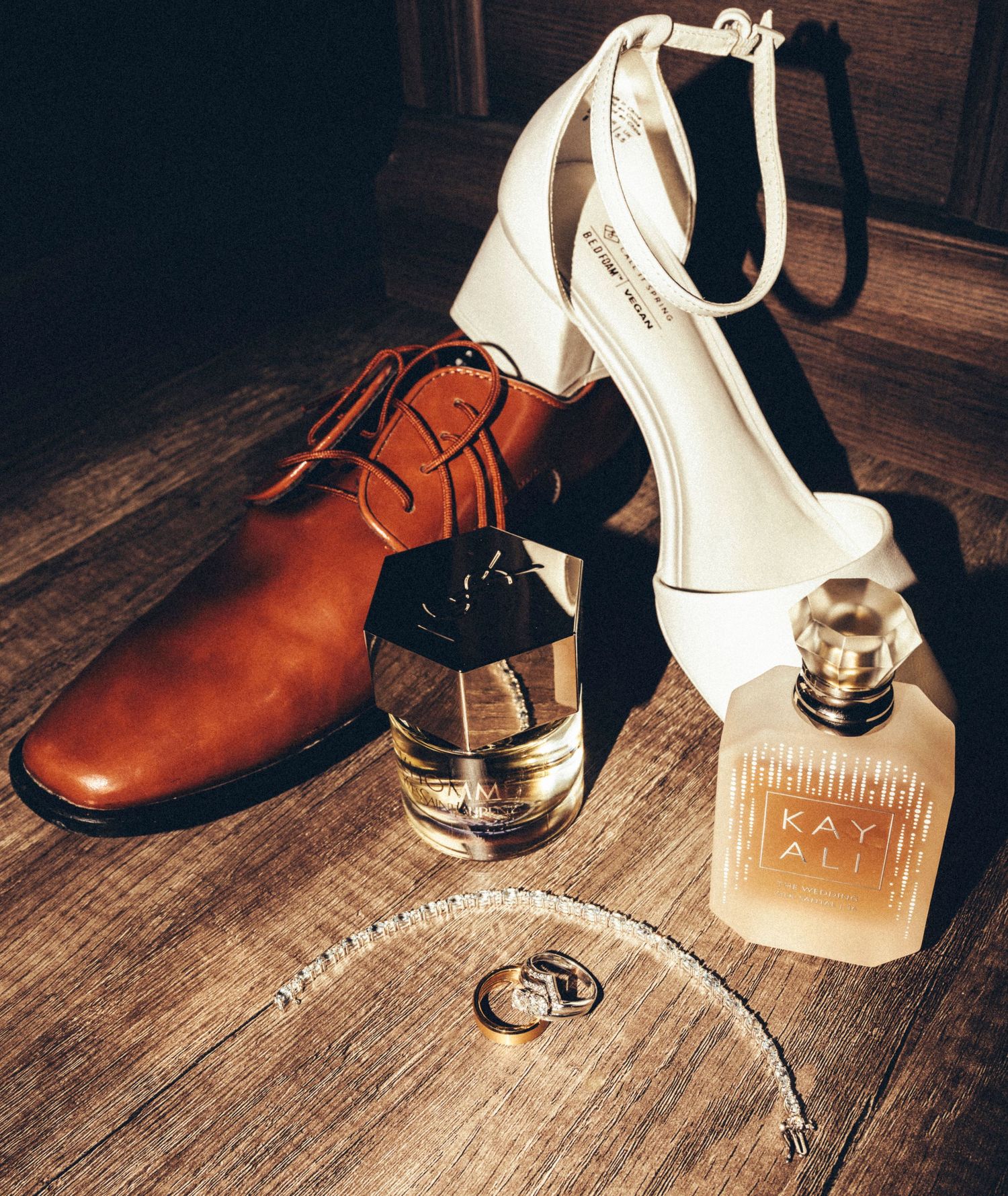 Wedding flat lay with brown oxford shoe, white heeled sandal, perfume bottles, and gold rings on wood.