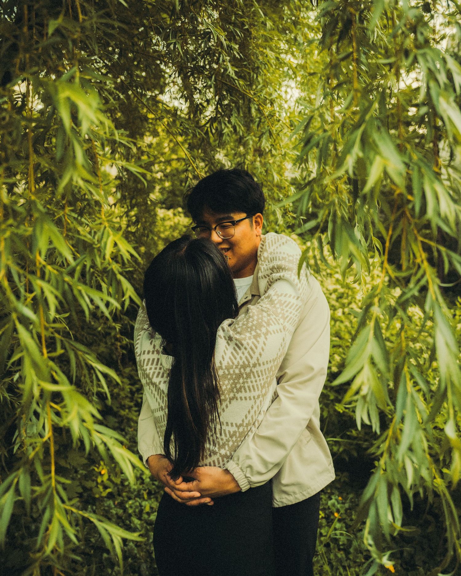 Couple embracing and smiling beneath weeping willow branches in a lush green outdoor setting.