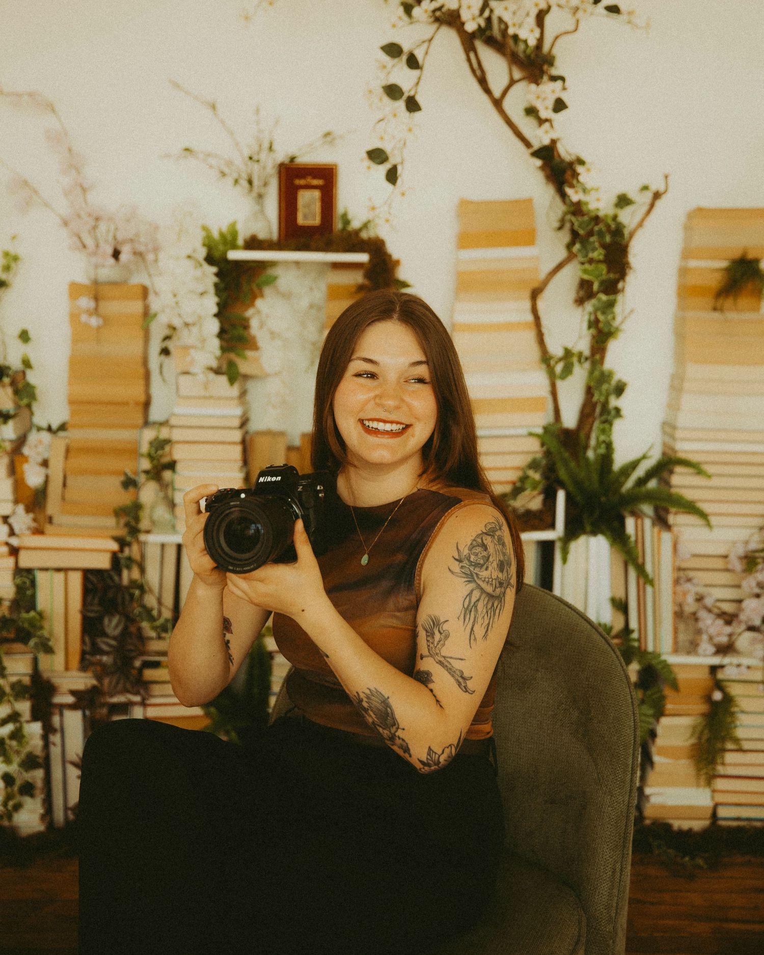 Smiling female photographer holding a Nikon camera seated among stacked books and floral decor.