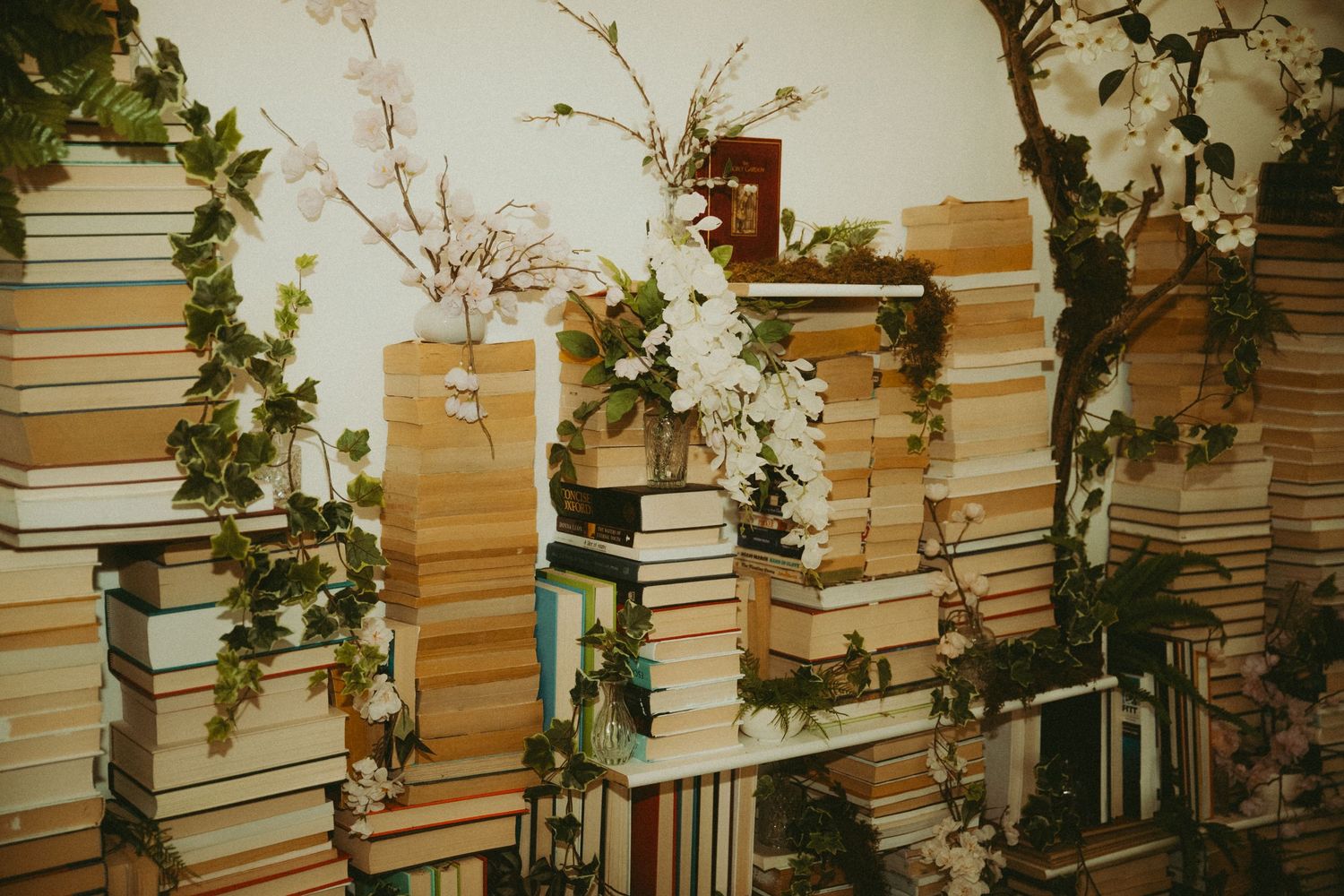 Decorative wall of stacked books adorned with white flowering branches and green ivy vines.