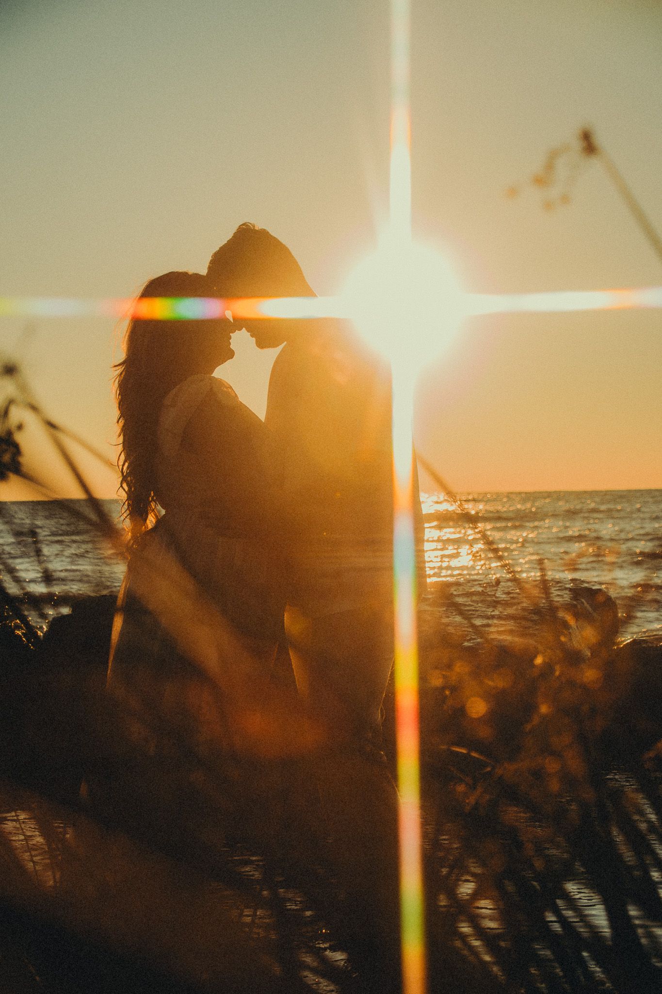 Romantic couple silhouetted against golden sunset with lens flare at ocean beach.