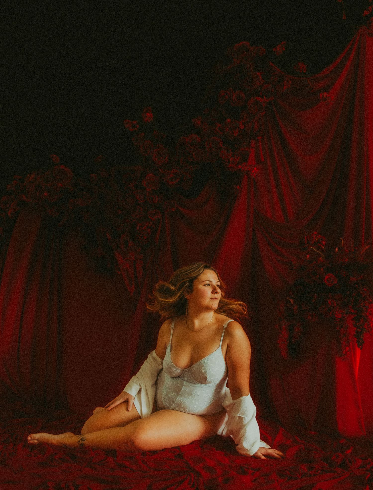 Woman in white lace lingerie and sheer robe seated on floor surrounded by red velvet drapes and dark florals.