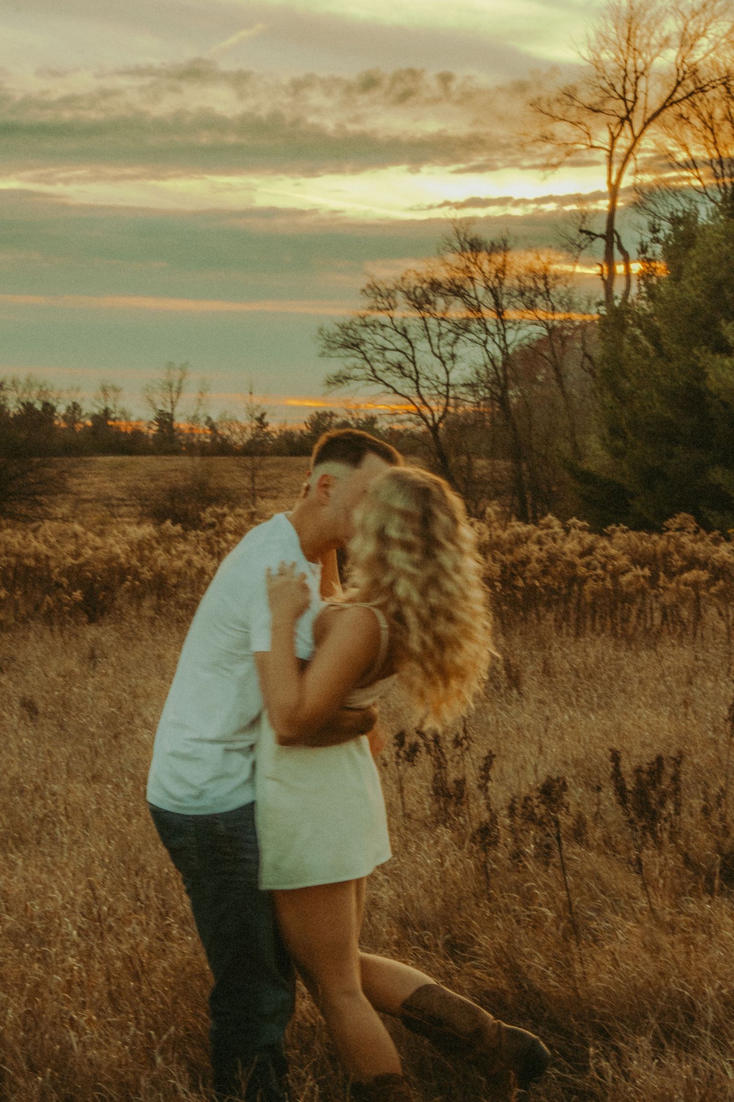 Couple sharing a kiss at sunset in a golden meadow, woman in white mini dress and cowboy boots.