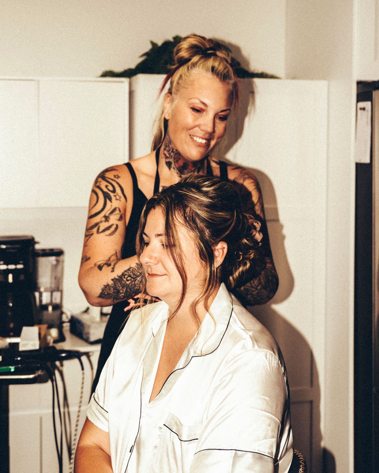 Heavily tattooed hairstylist smiling while styling bride's updo hair before wedding ceremony.