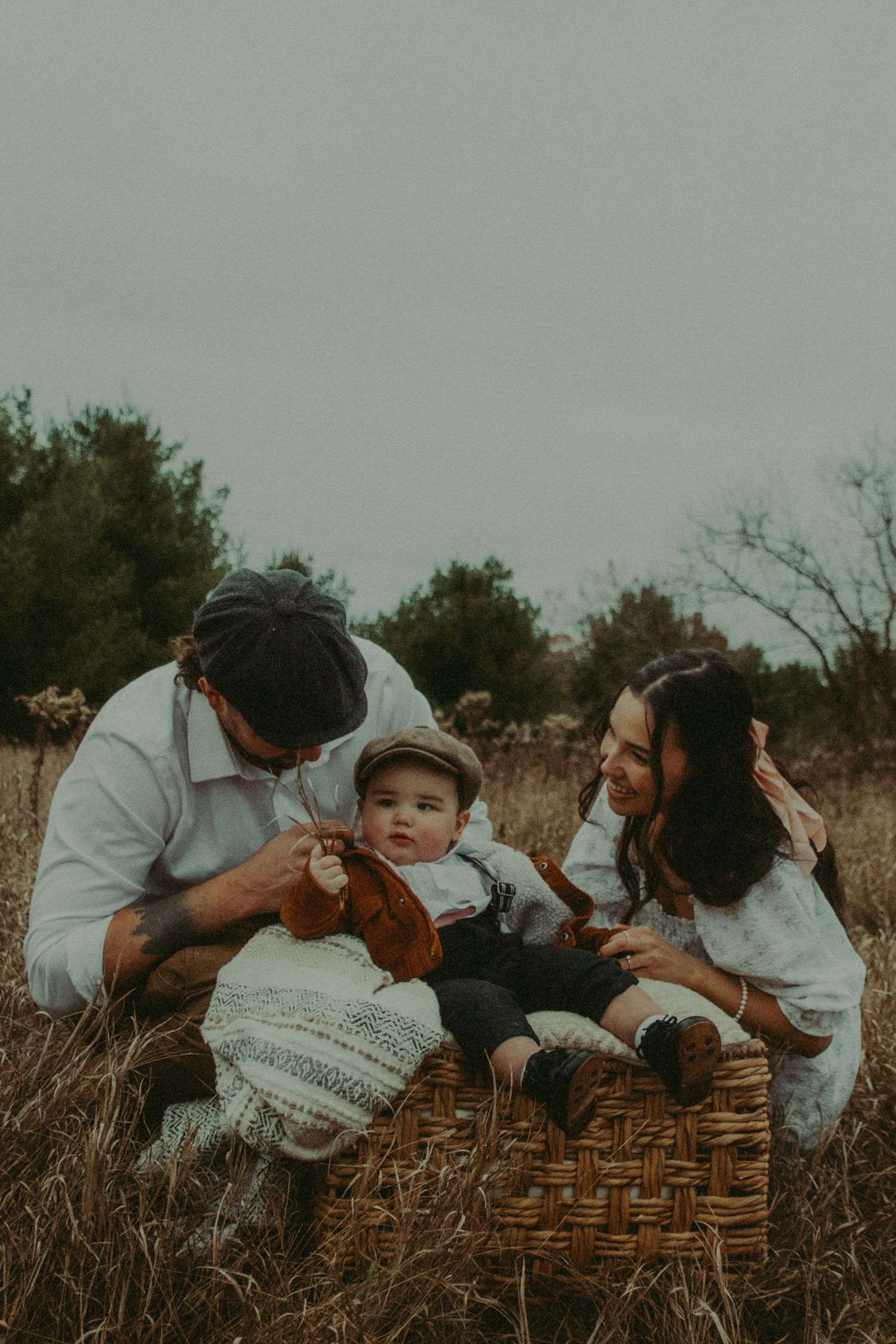 Parents playing with baby sitting in wicker basket in an open field on overcast day.