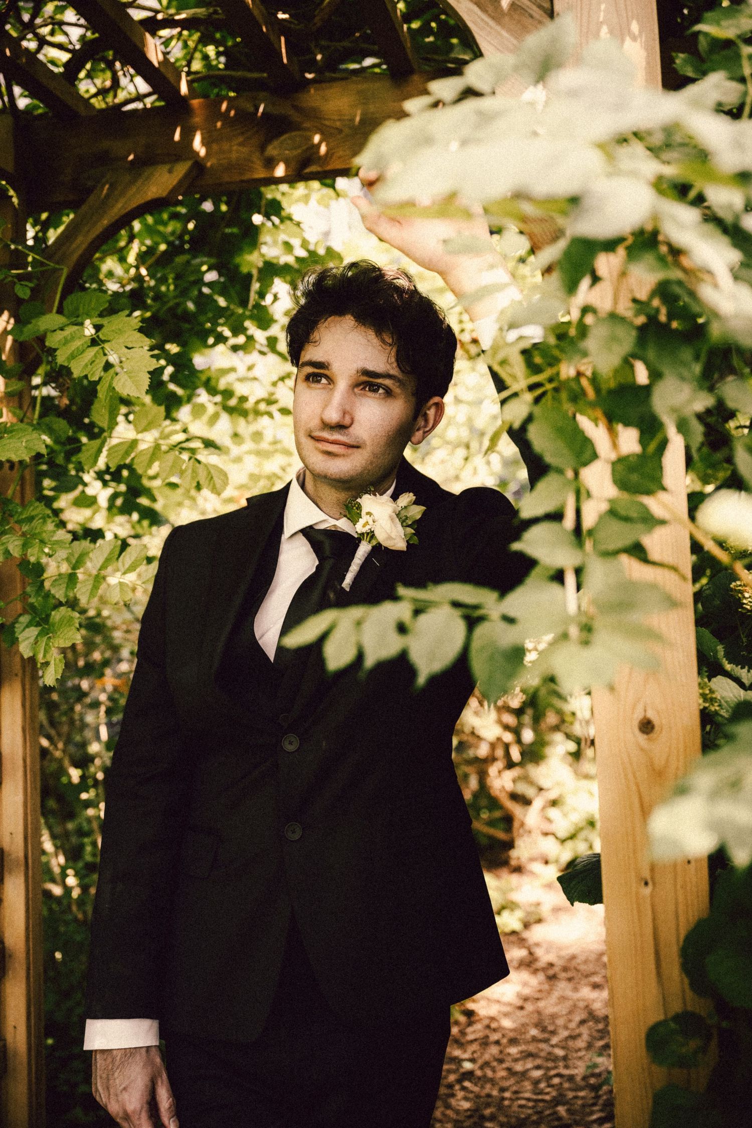 Handsome groom in black tuxedo with boutonniere stands beneath a wooden arbor covered in lush green leaves.
