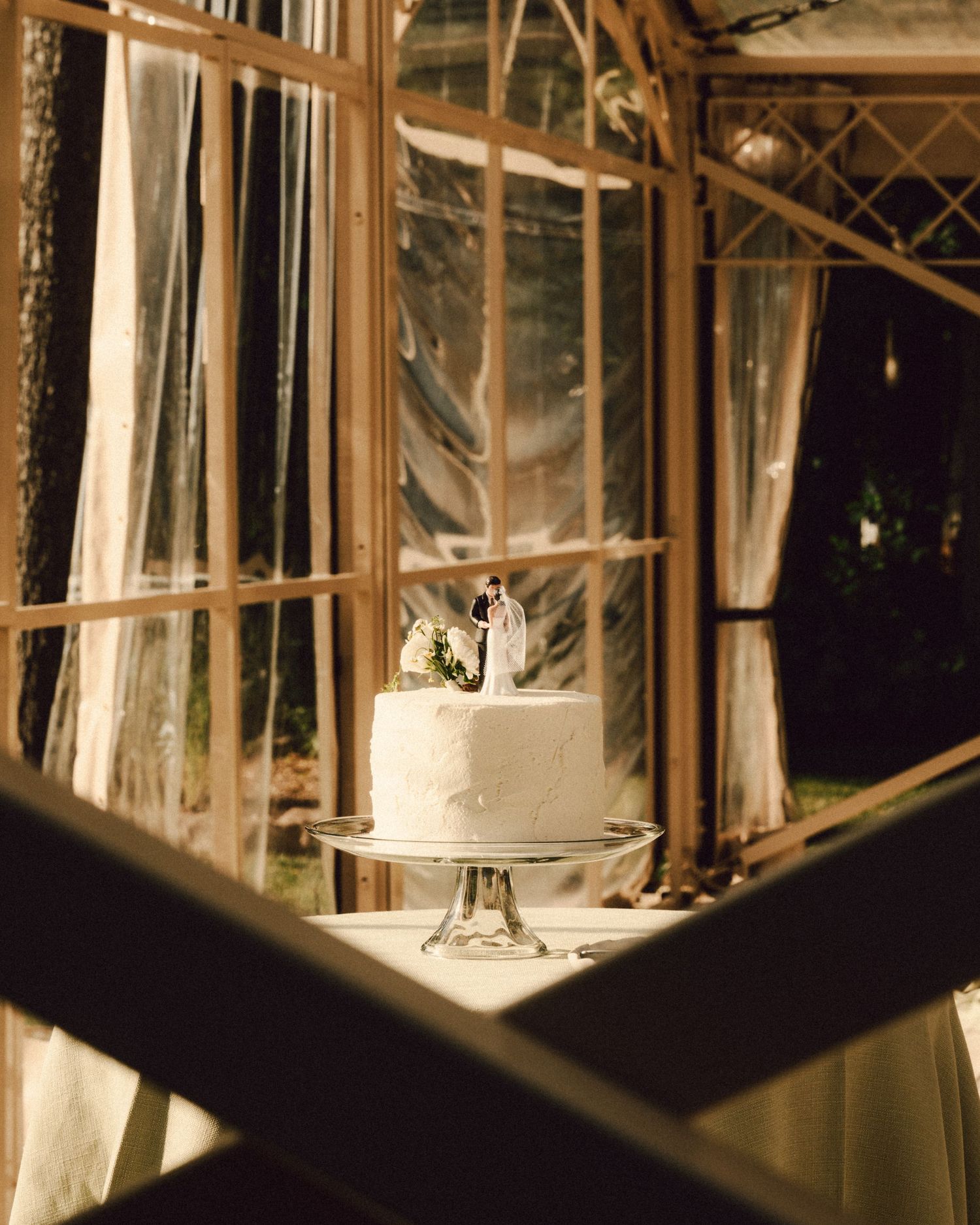 Simple two-tier white wedding cake with bride and groom topper displayed in warmly lit glass greenhouse venue.
