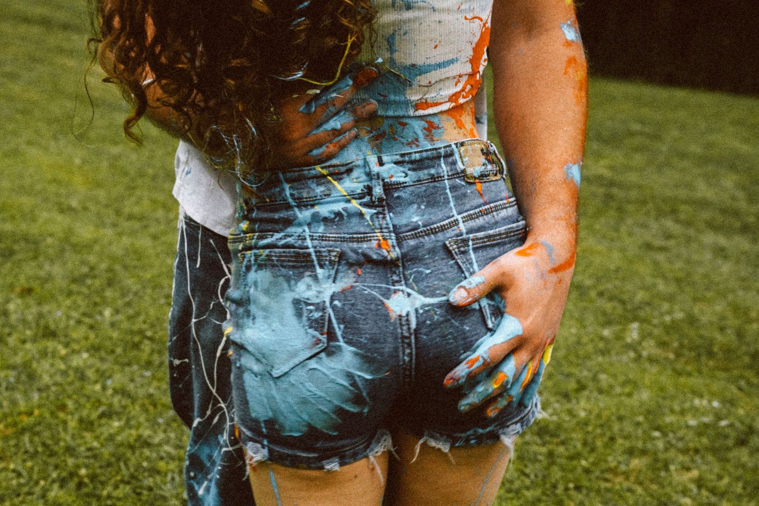Back view of woman in paint-splattered denim shorts with colorful handprints on grass.