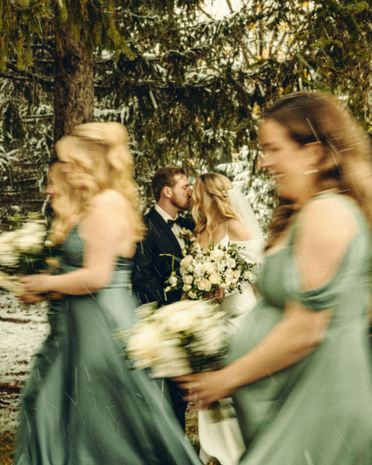 Bride and groom kissing while bridesmaids in sage green dresses walk past in motion blur.