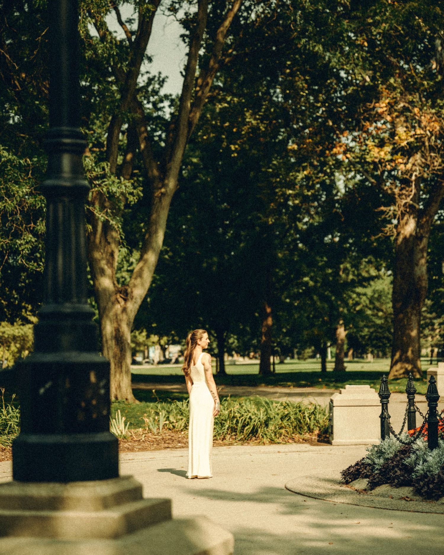 Bride in elegant white gown standing alone on park pathway surrounded by lush green trees.