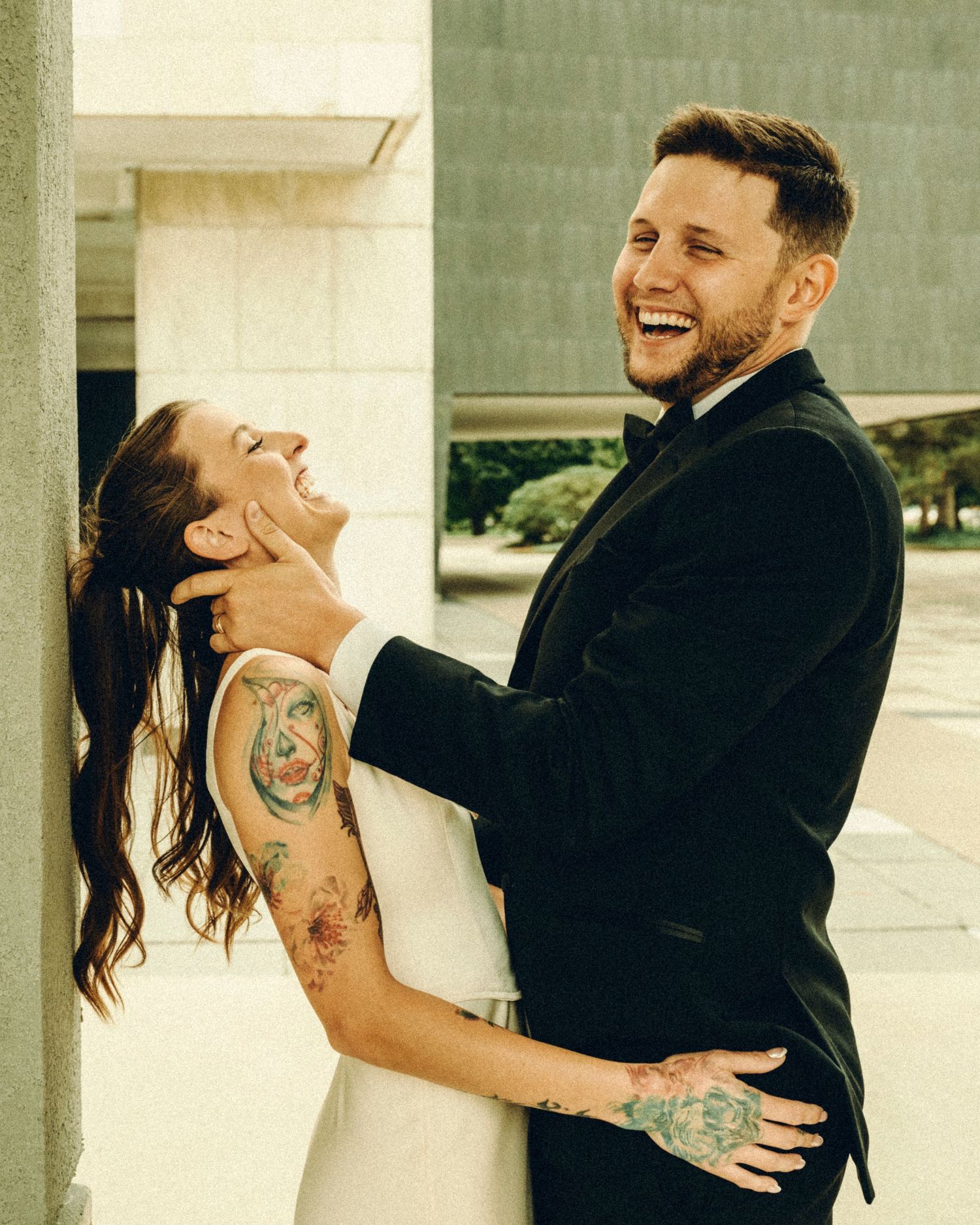 Laughing bride and groom in wedding attire sharing a joyful embrace outdoors, both with tattoos.