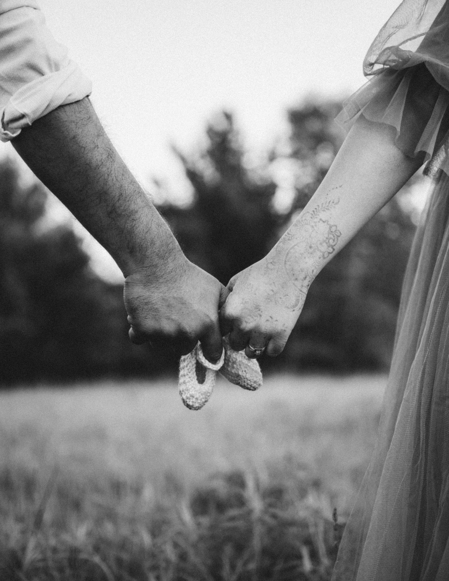 Black and white photo of couple holding tiny baby shoes together in an outdoor field setting.