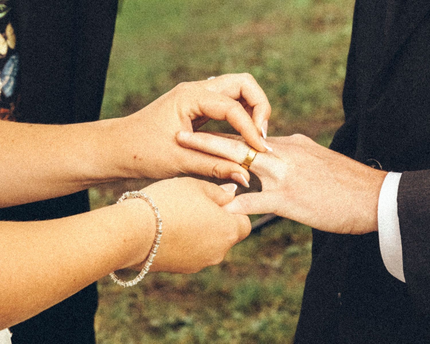 Close-up of groom placing gold wedding ring on brides finger during outdoor ceremony.