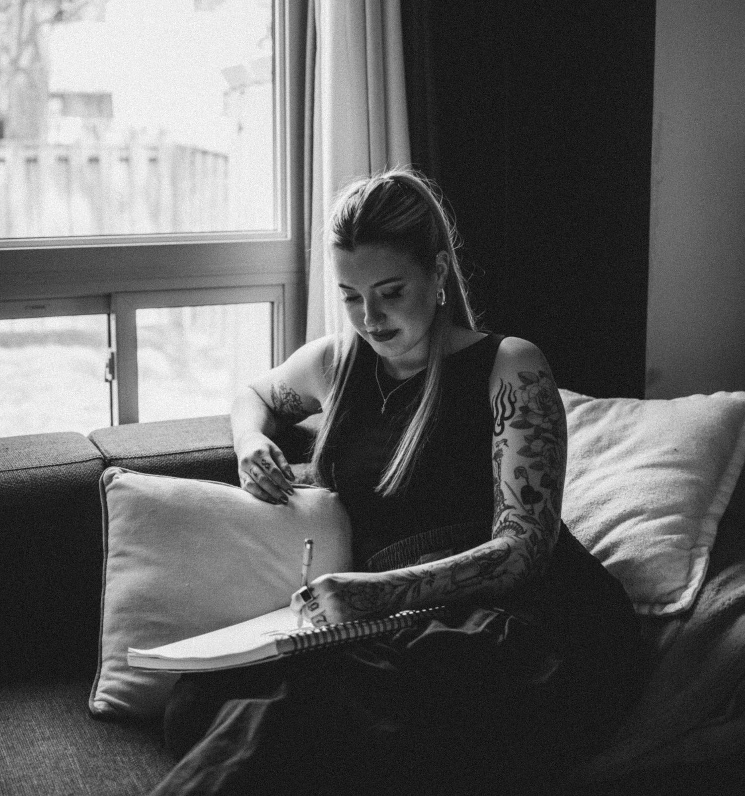 Black and white photo of tattooed woman writing in notebook on couch near window indoors.