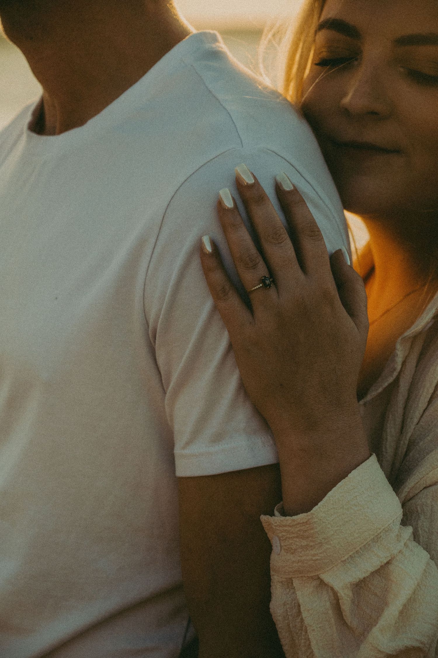 Woman with white nails and engagement ring resting hand on man's white t-shirt at sunset.