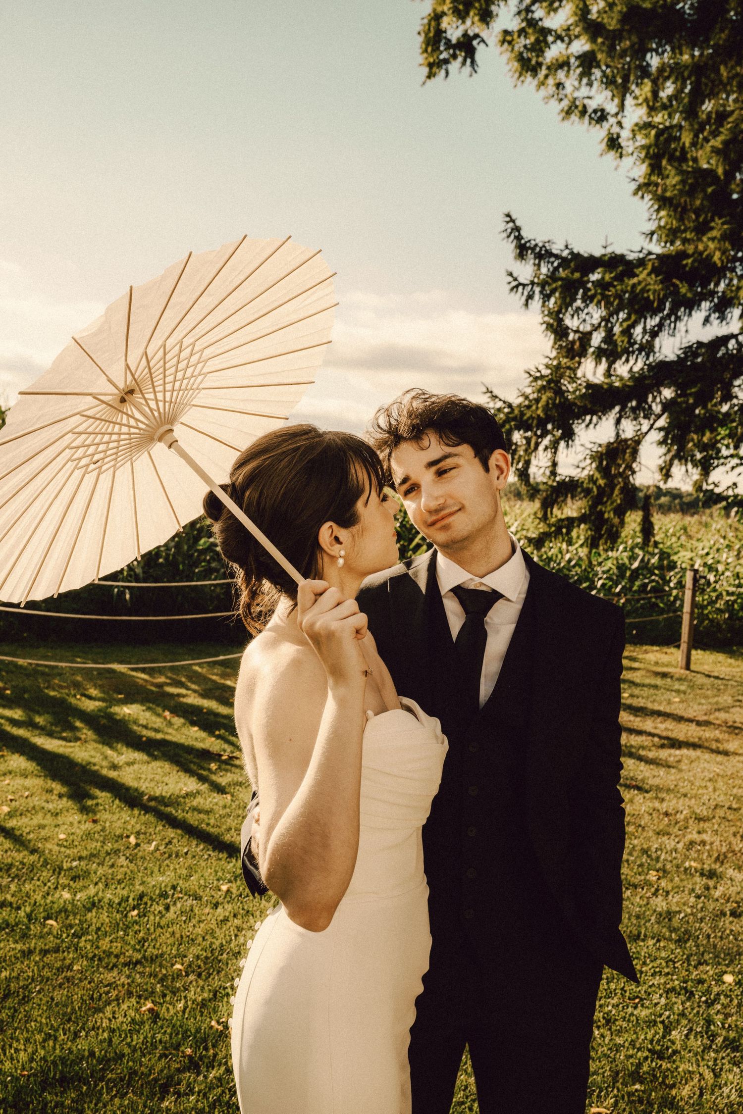 Bride in strapless gown and groom in black suit share an intimate moment with white parasol outdoors.
