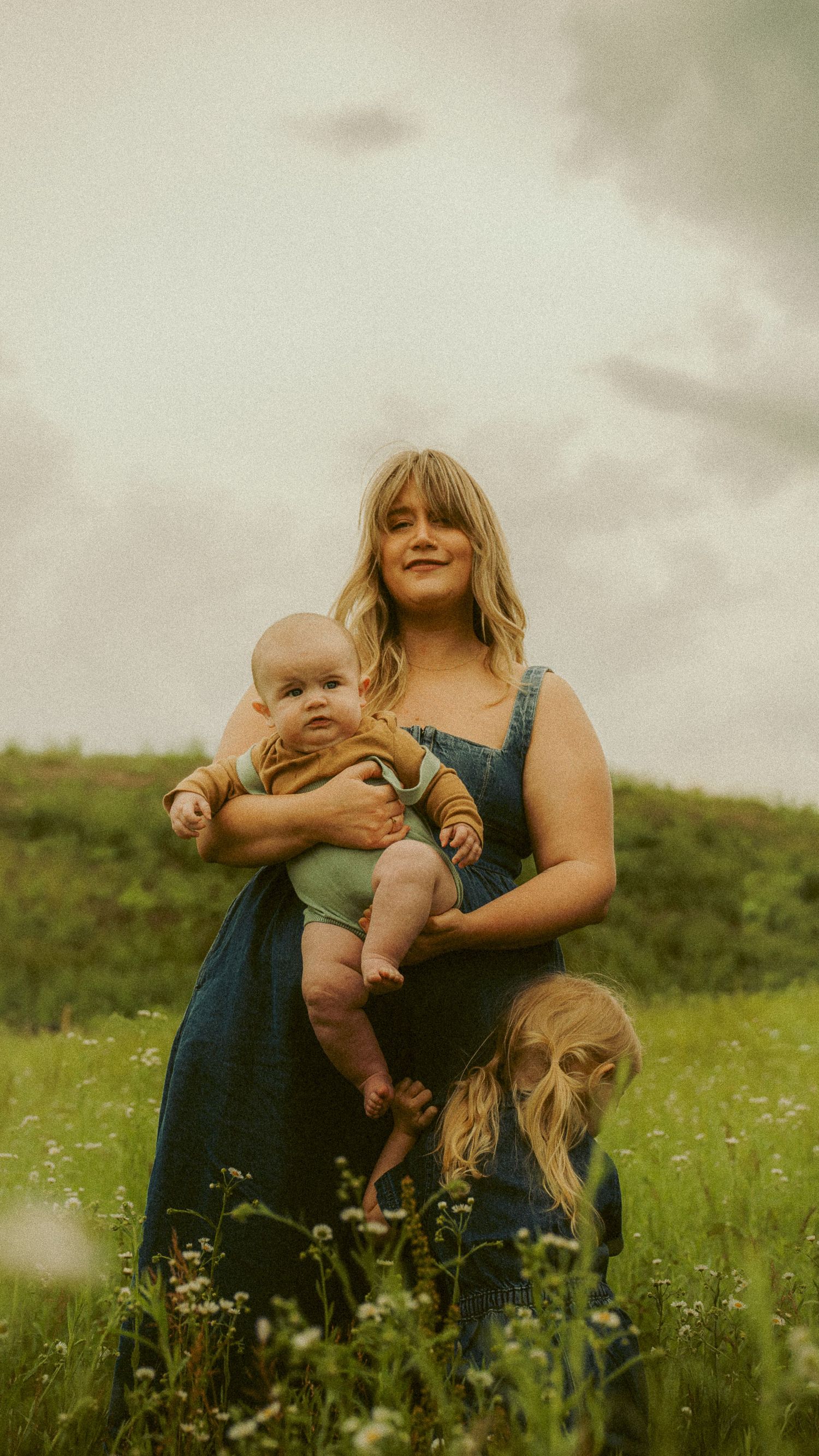 Mother in blue denim dress holding baby in mustard outfit while standing in wildflower field under cloudy sky.