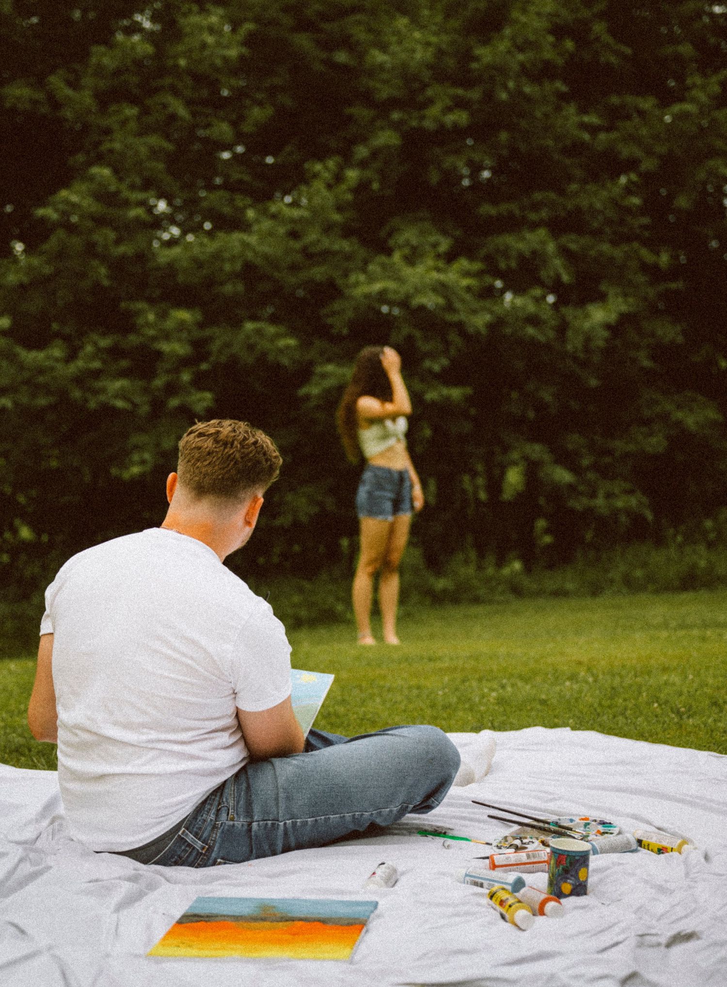 Man sitting cross-legged on blanket painting while woman stands near trees in sunny park.