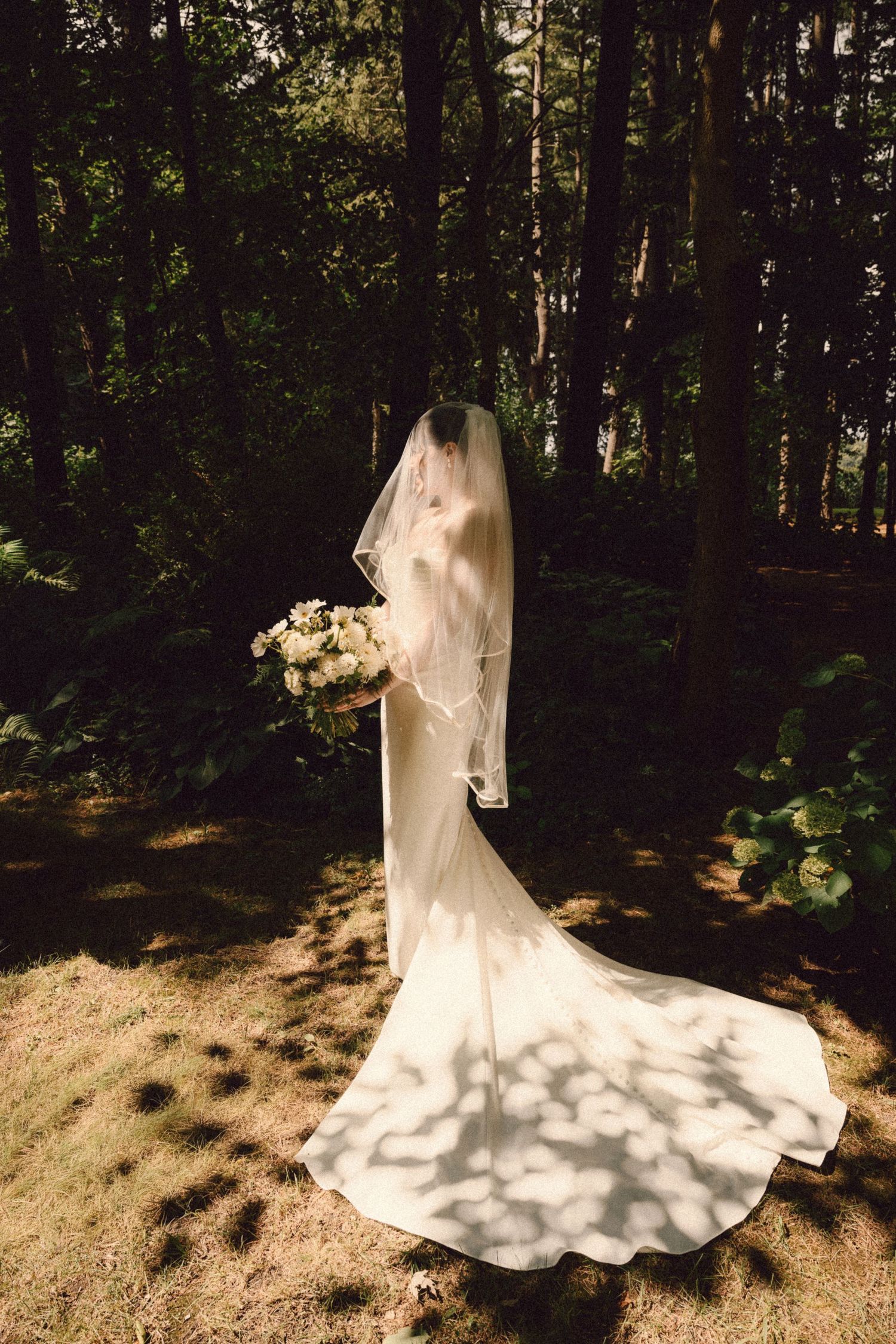 Bride in flowing white gown and veil stands in dappled sunlight among tall trees holding bouquet.