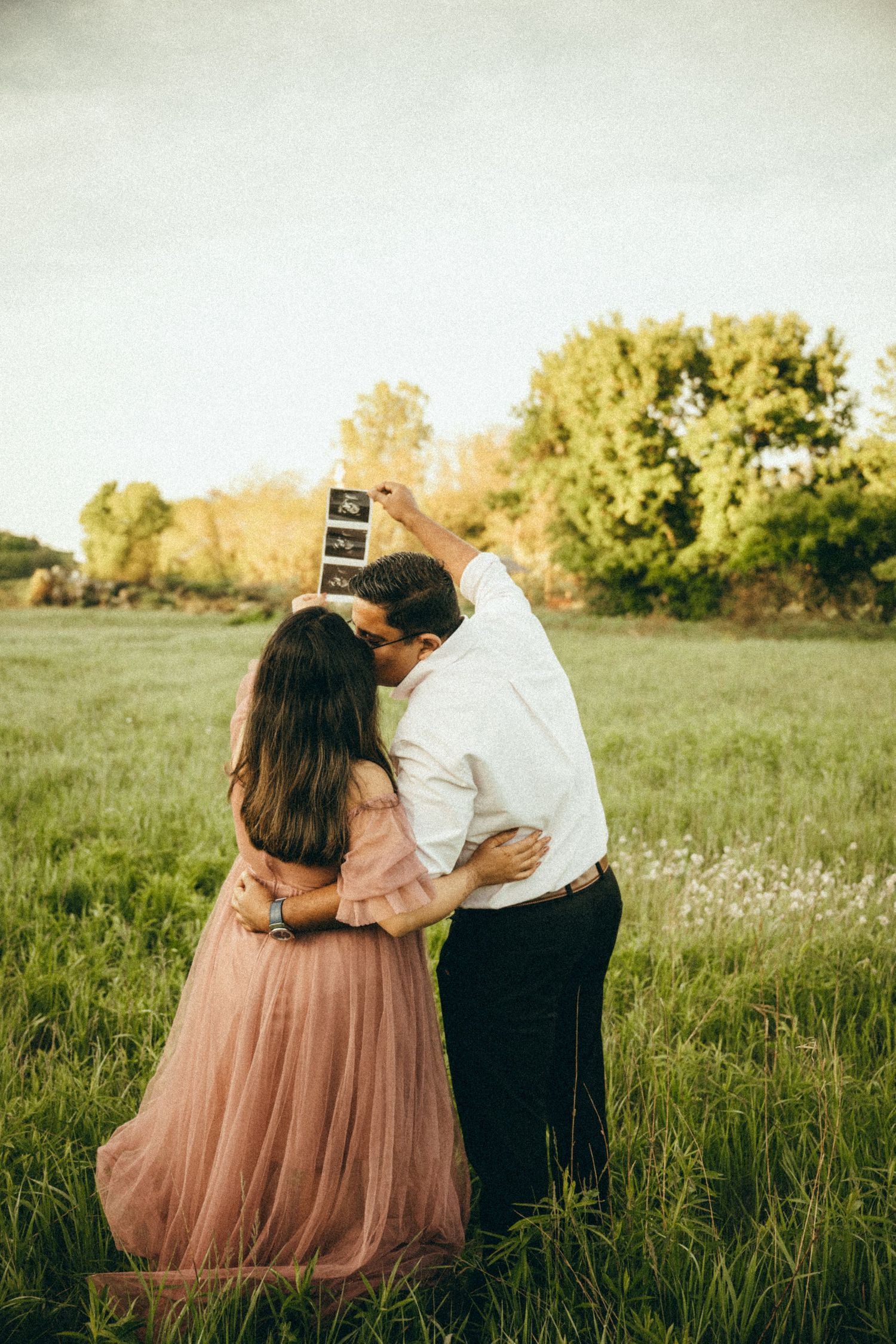 Expecting couple kissing while holding ultrasound photos up in a scenic green field at sunset.